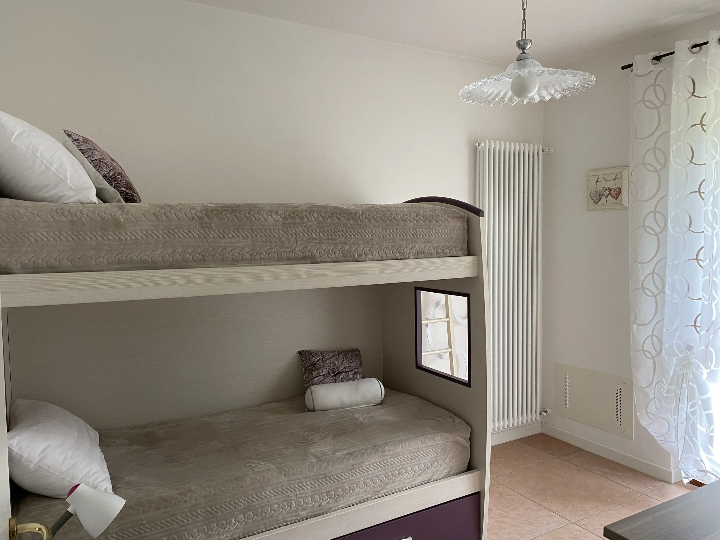 A room with white walls, a white radiator, a window with patterned white curtains, a ceiling lamp with a glass shade, and a bunk bed with beige mattresses, pillows, and a small mirror attached to the side of the lower bunk.