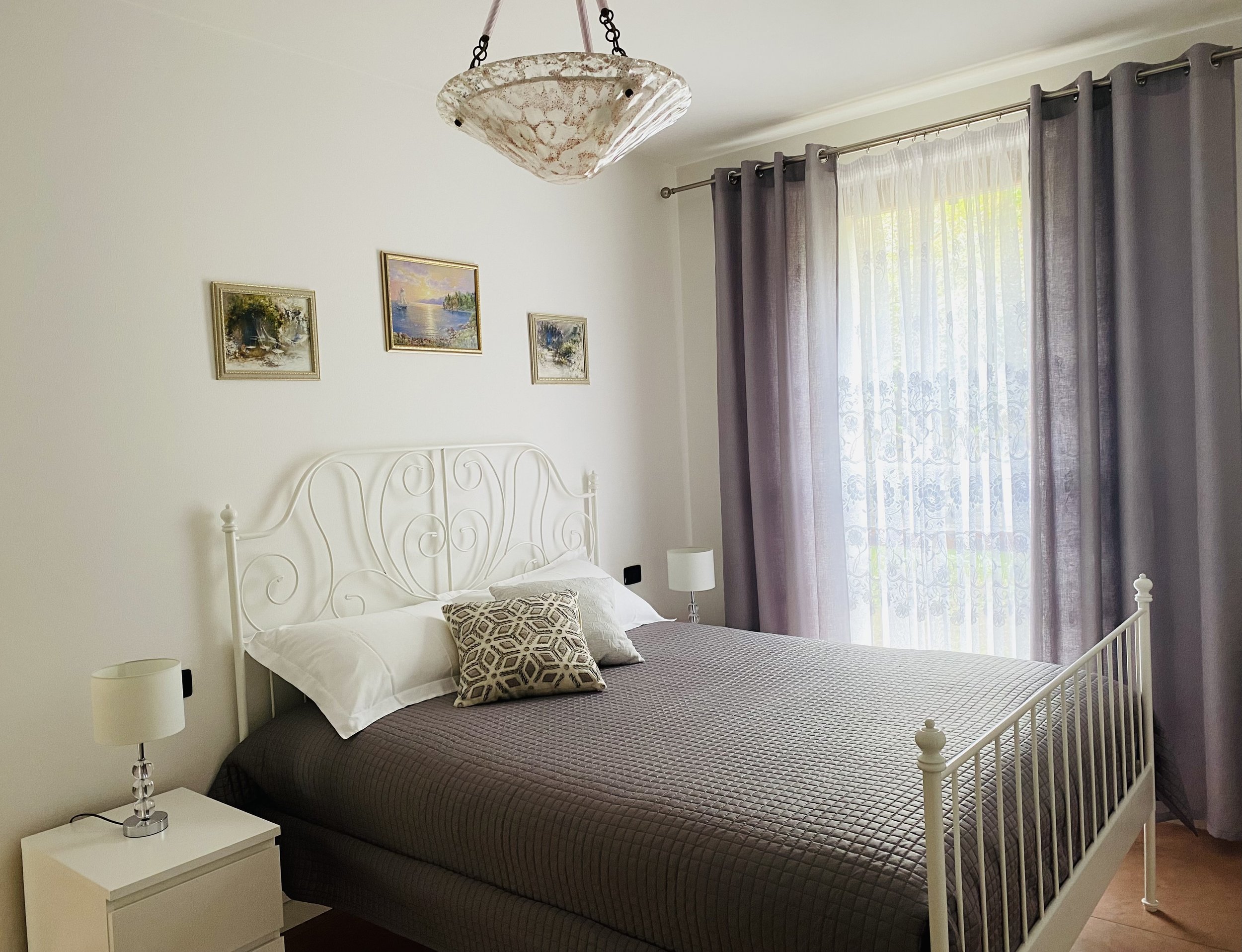 A bedroom with a white metal bed frame, beige and gray bedding, and decorative pillows. There are two white nightstands with lamps on each side of the bed. The wall above the bed has three framed landscape paintings. A large window with sheer white c