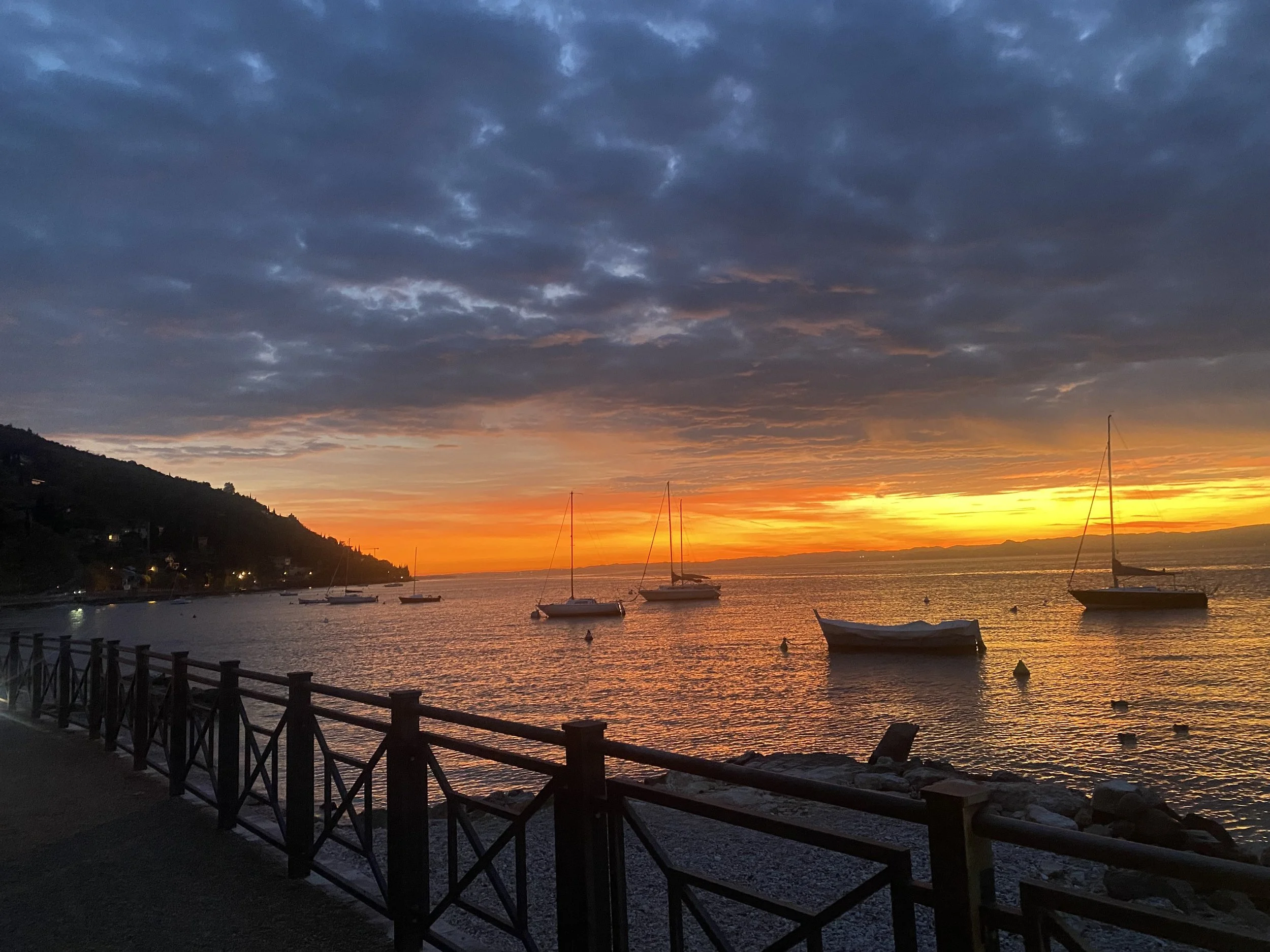Sunset over a harbor with sailboats, calm water, and a mountain on the left, with a cloudy sky.