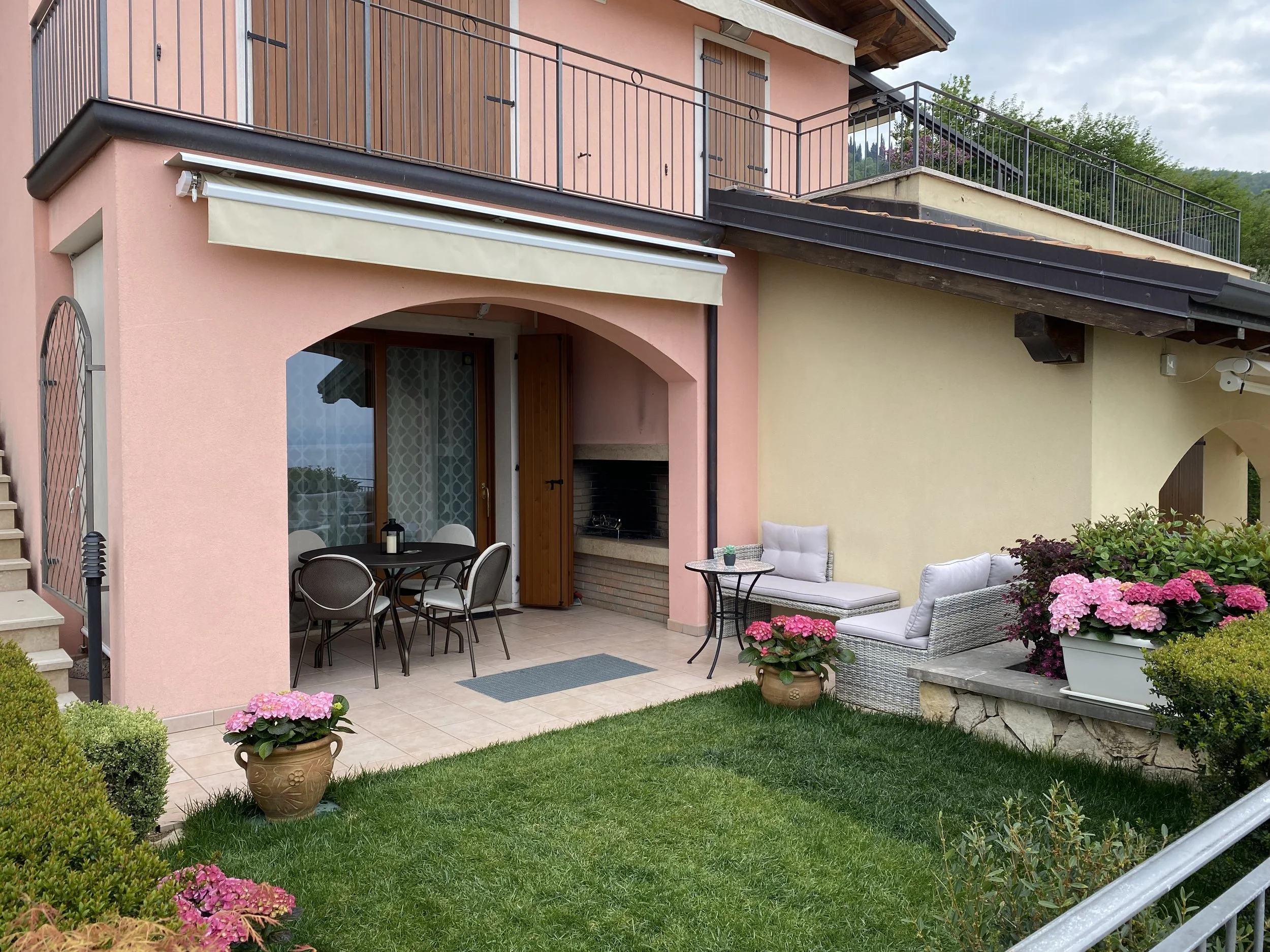Exterior view of a two-story house with a pink and yellow facade, outdoor patio with chairs and a table, potted pink flowers, and outdoor furniture, with a small green lawn and lush greenery in the background.