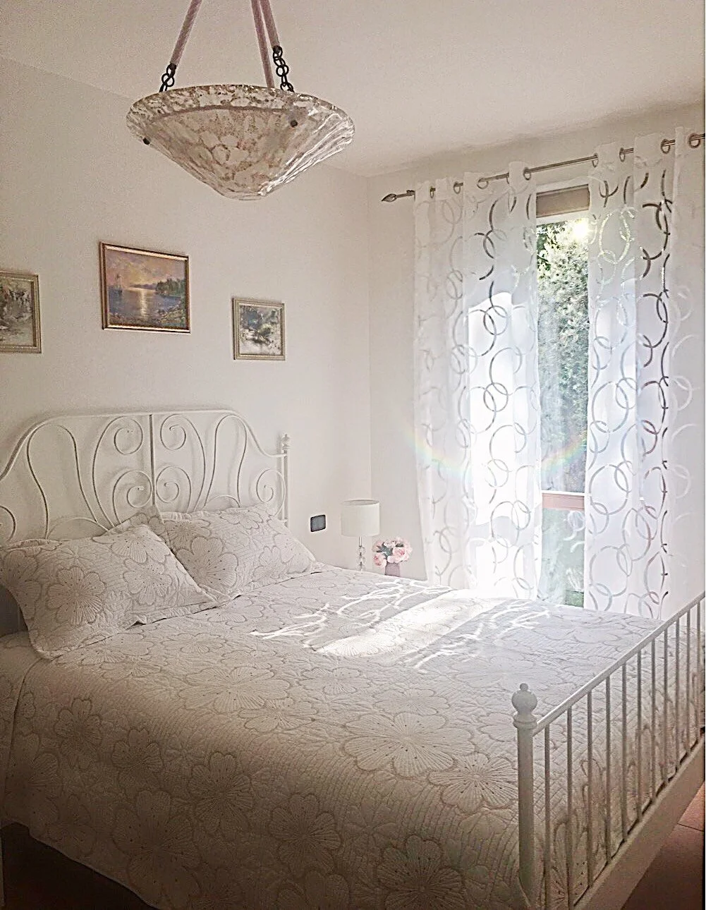 A bedroom with white walls, a white metal bed frame, floral bedding, and matching pillows. There are three framed landscape paintings on the wall above the bed. A small nightstand with a white lamp and flowers is next to the bed. White patterned curt