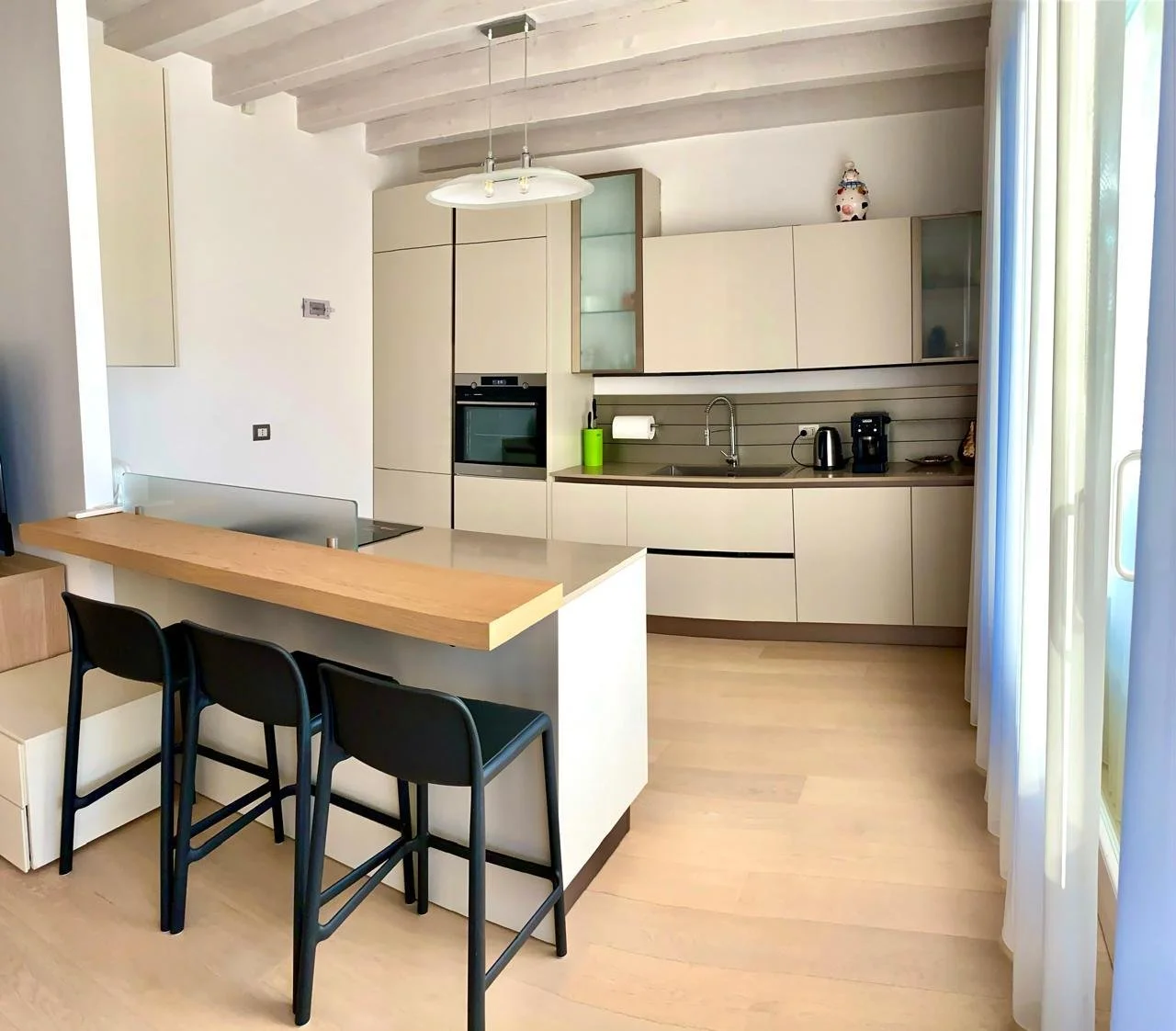 Modern kitchen with light-colored cabinets, a sink, a coffee maker, and a built-in oven. A wooden breakfast bar with black chairs is in the foreground, and large windows with white curtains on the right.