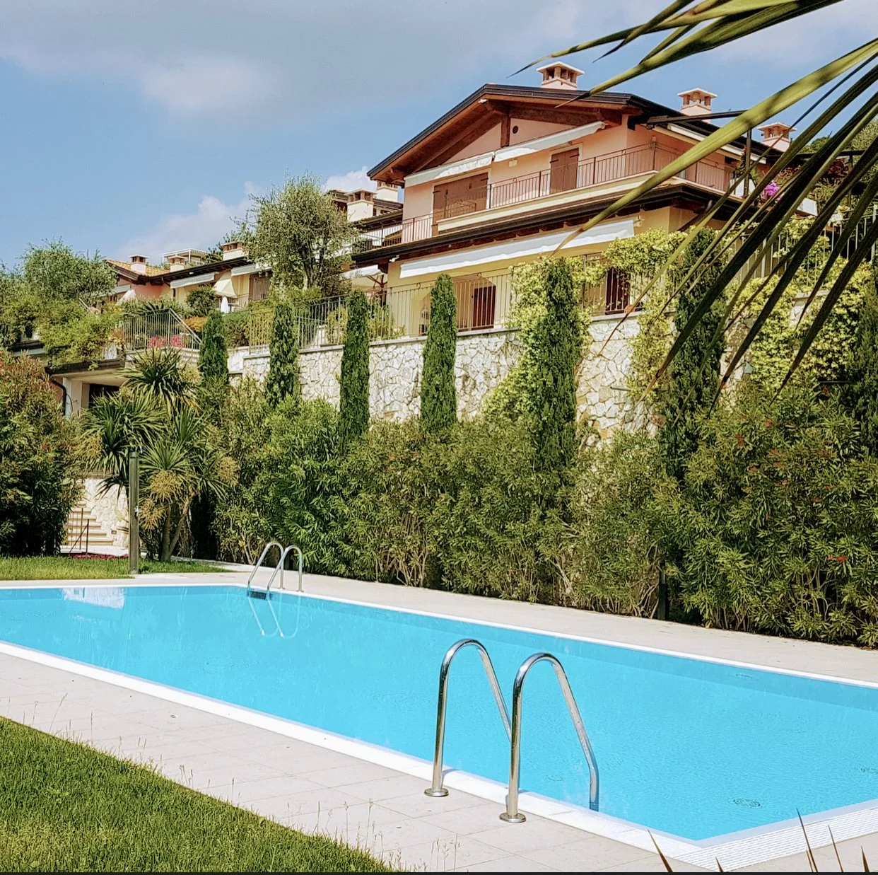 Swimming pool with clear blue water in a backyard, surrounded by green grass, bushes, and tall trees, with multi-story houses with balconies and a stone wall in the background.