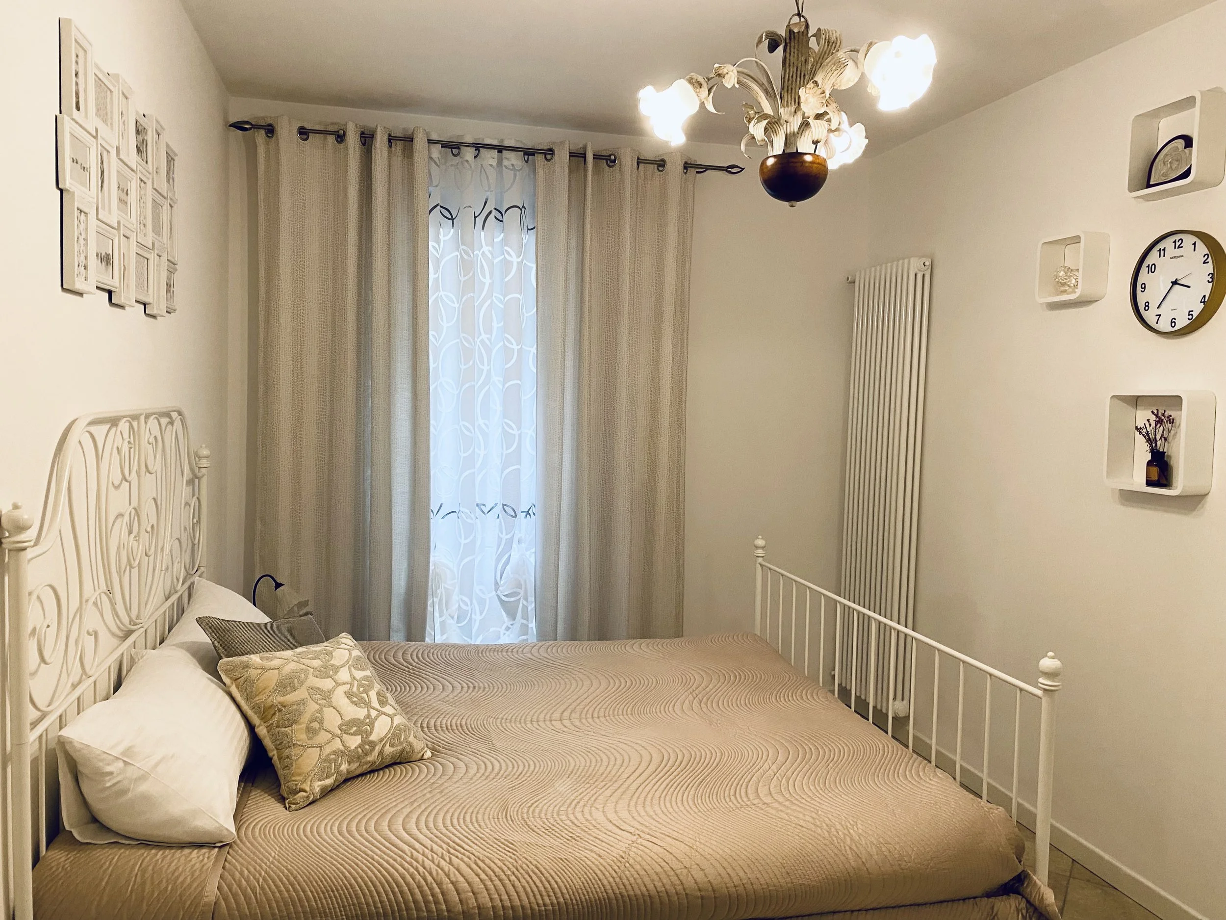 A bedroom with a white metal bed frame, beige bedding, and pillows. There are framed pictures on the left wall, beige curtains and sheer curtains on the window, and a chandelier hanging from the ceiling. The right wall has a radiator, a clock, and wh