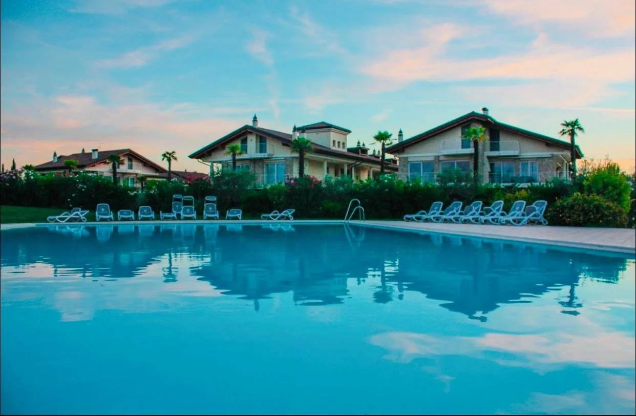 A swimming pool surrounded by lounge chairs with residential houses and palm trees in the background during sunset or sunrise.