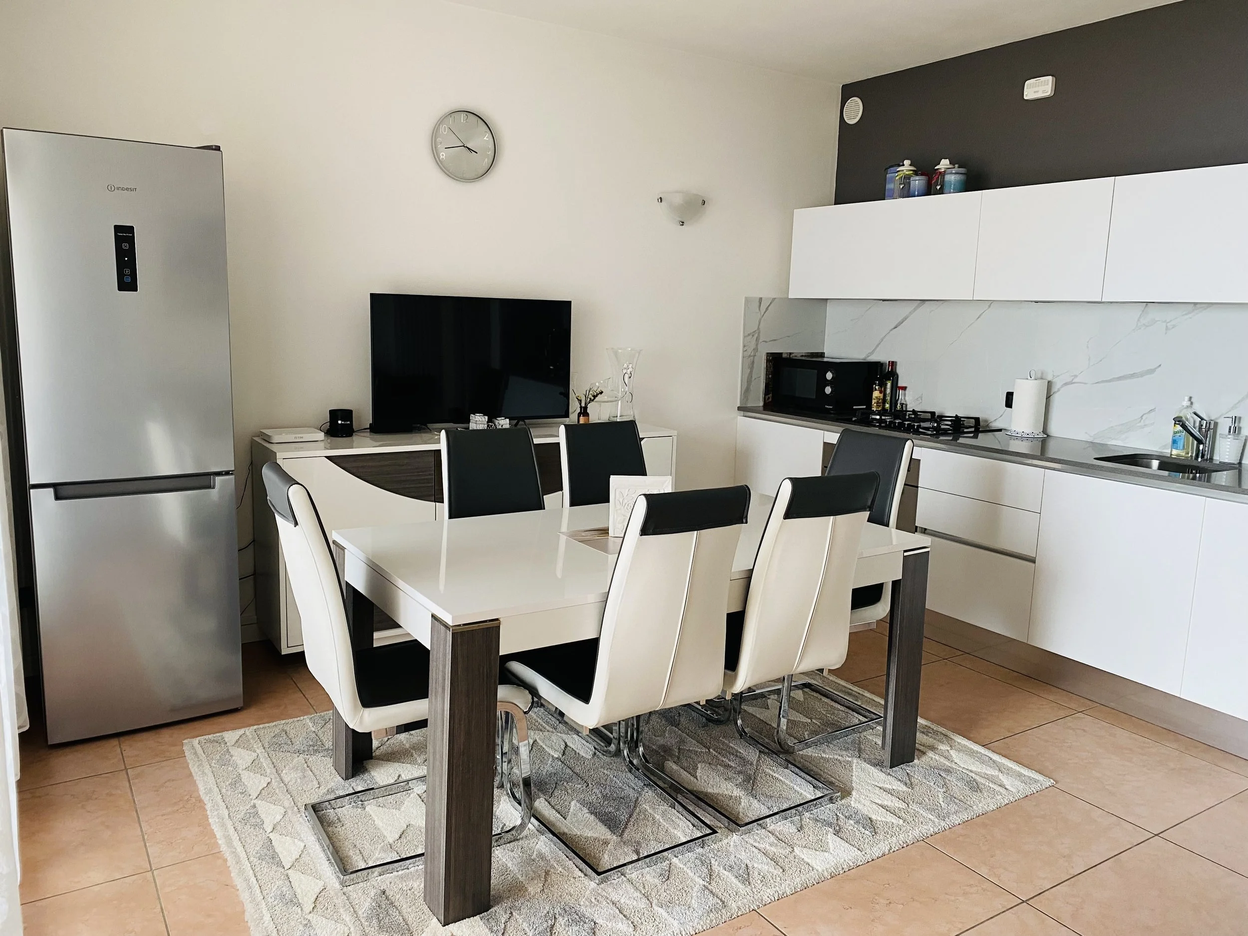 Modern kitchen with a white dining table and six chairs, a black refrigerator, a TV on a white console, and a white kitchen counter with appliances and utensils.