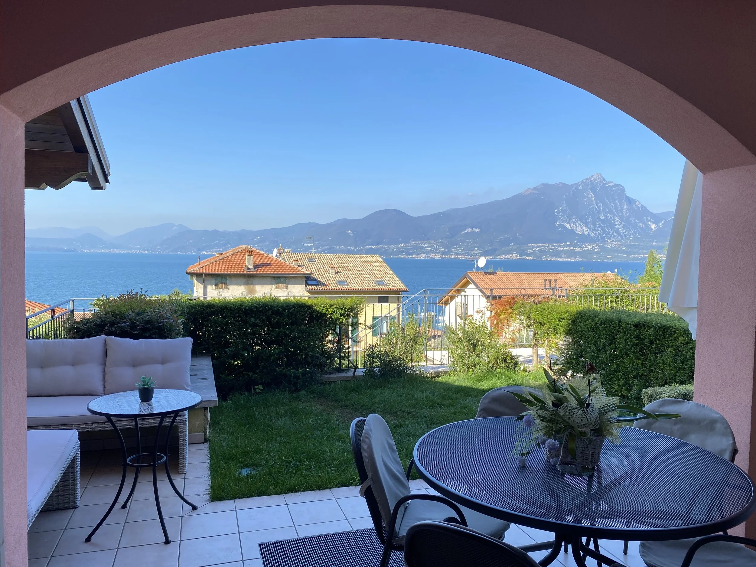 View of a lake with mountains in the background, seen from a patio with outdoor furniture, including a round table with a flower arrangement, a sofa, and a small side table, under an arched pink stucco ceiling.
