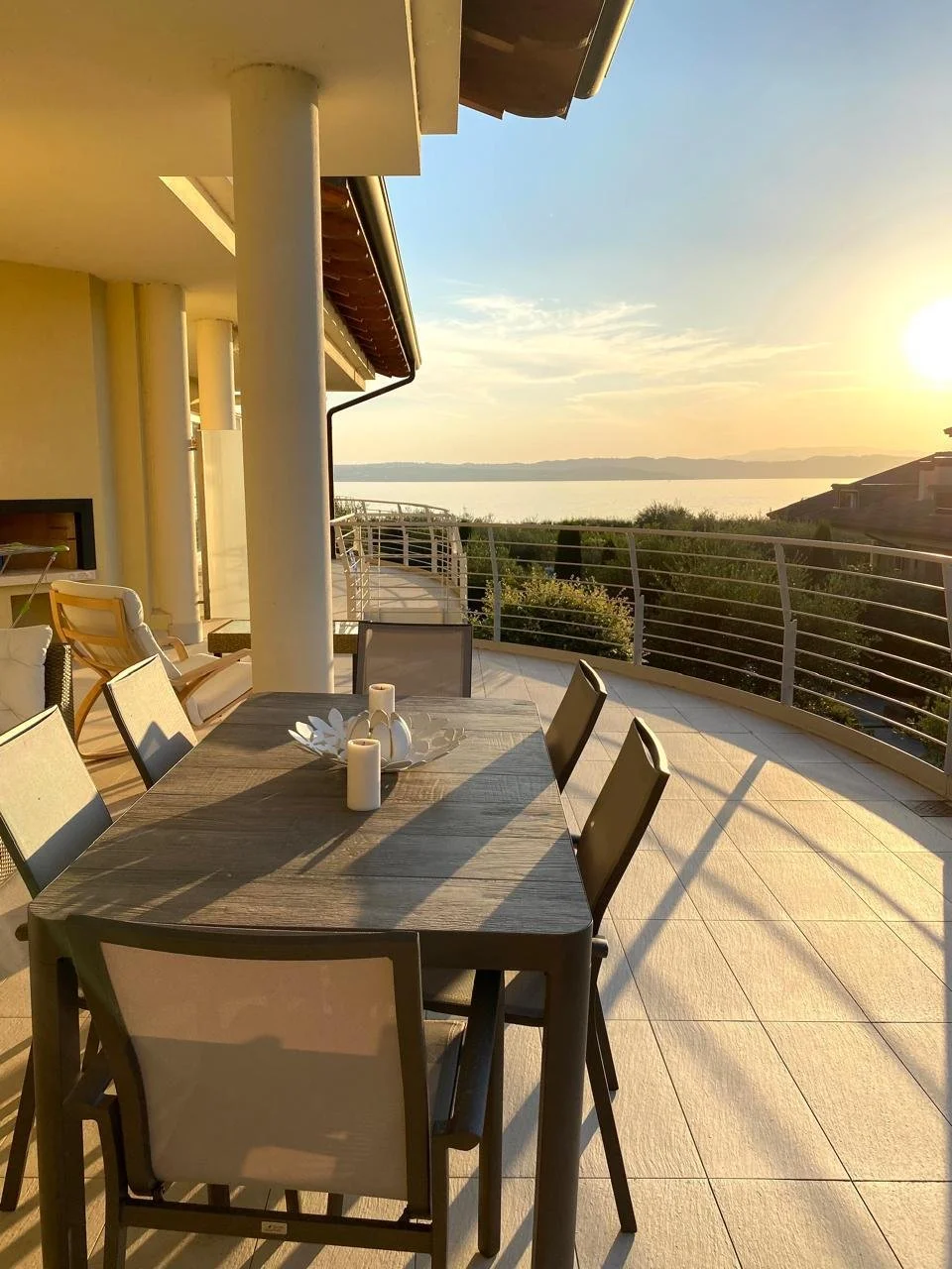 Outdoor balcony with a wooden dining table and six chairs, overlooking a body of water at sunset, with a partly cloudy sky.