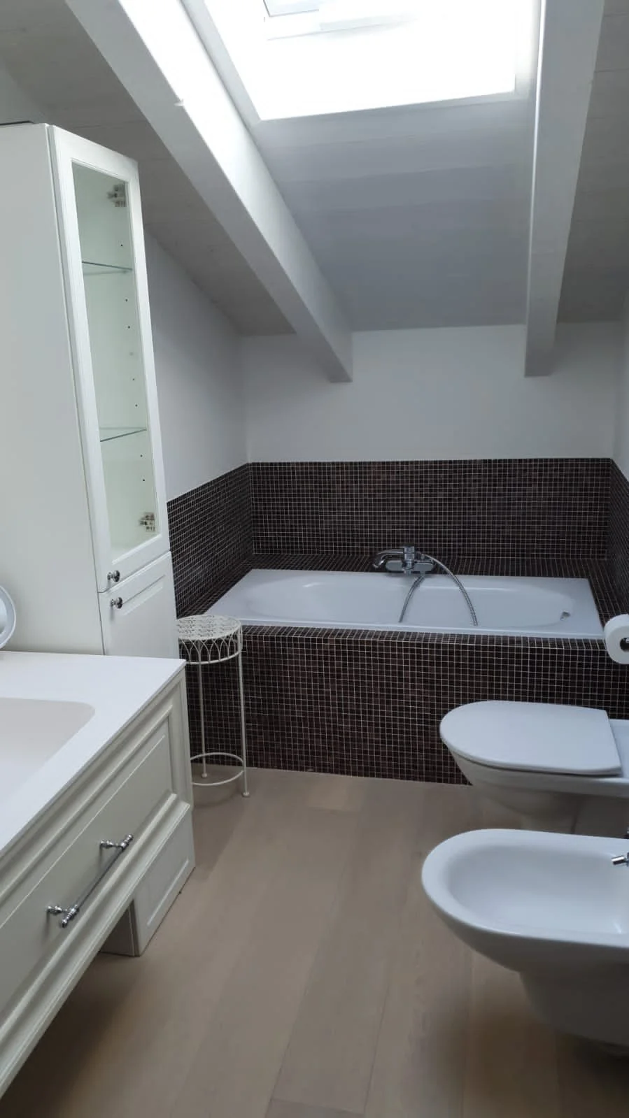 Bathroom with a skylight, bathtub with black mosaic tiles, white vanity, toilet, bidet, and a white cabinet.