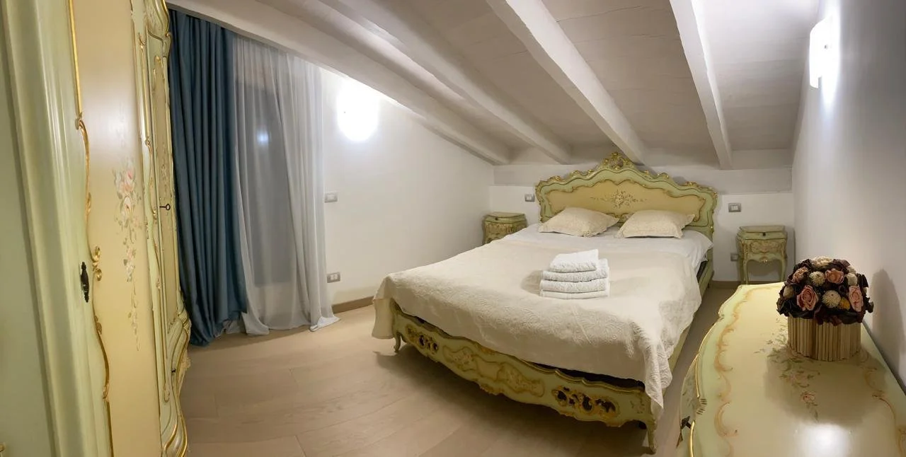 A bedroom with a bed, bedside tables, a wardrobe, and a vase of flowers. The room has sloped ceilings and is decorated in pastel green and cream colors.