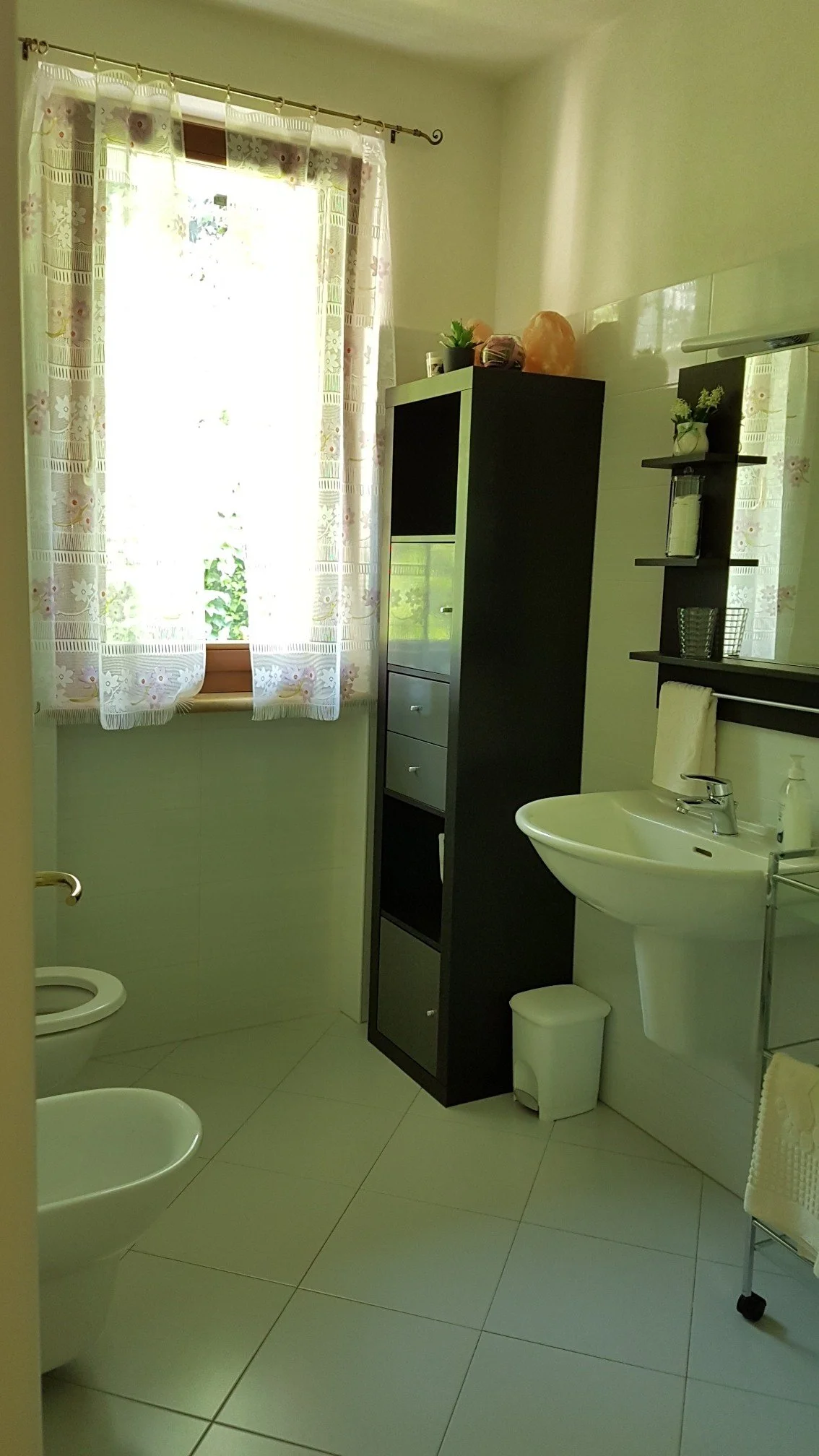 A bathroom with a window with floral curtains, a white sink, a black storage cabinet, a towel rack, and a bidet.