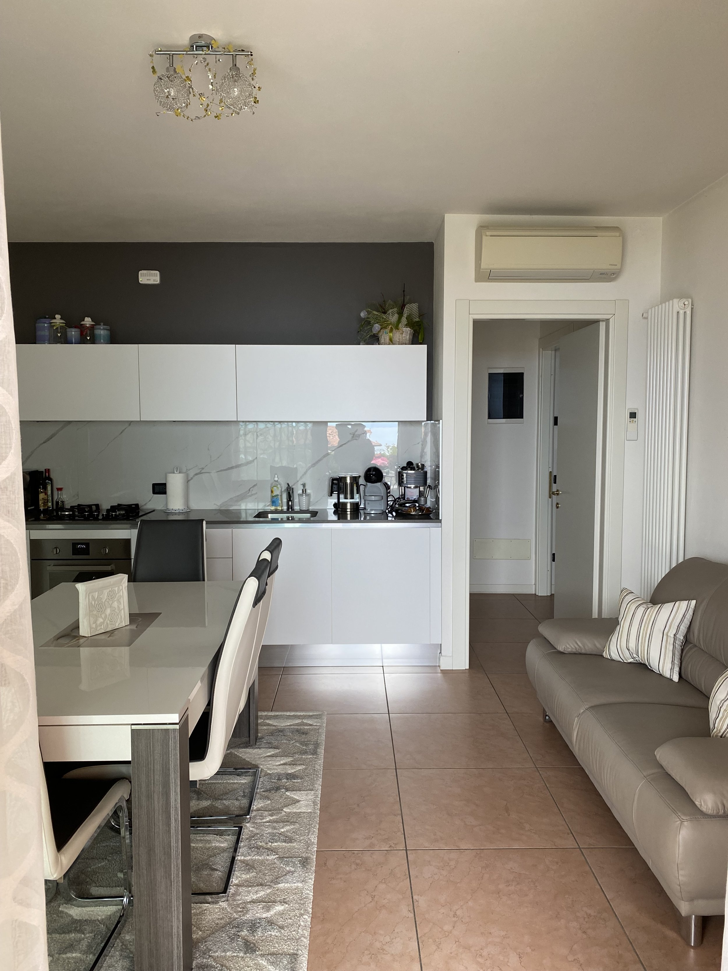 Living room and kitchen area with a tiled floor, white cabinets, dining table with chairs, gray sofa with striped pillows, and small appliances on the kitchen counter, including a coffee maker and toaster.