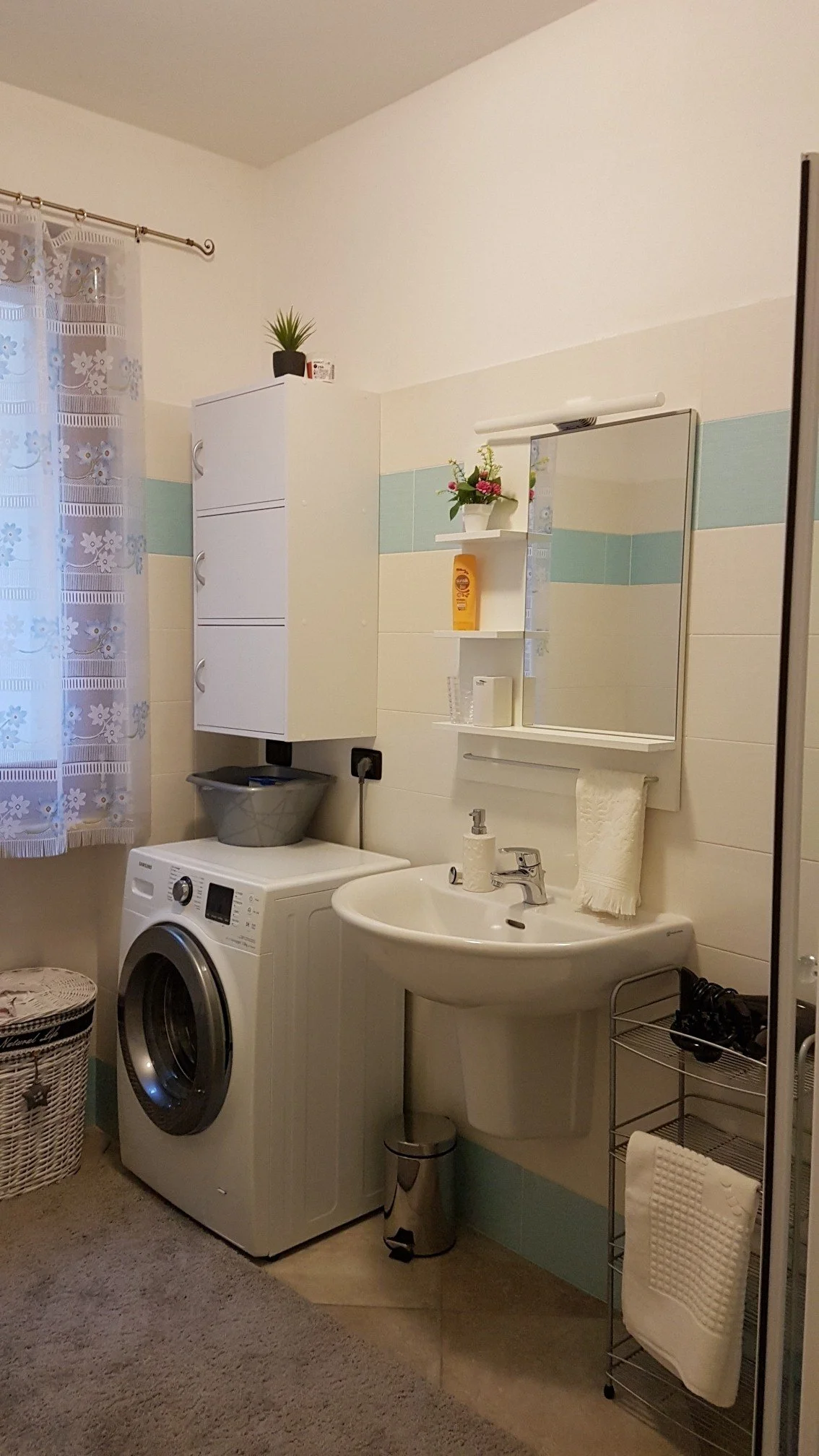 A bathroom with a laundry area featuring a washing machine, a small sink with a mirror above, shelves with decorative items and personal care products, a laundry basket, and a small potted plant on top of a cabinet. There's a window with lace curtain