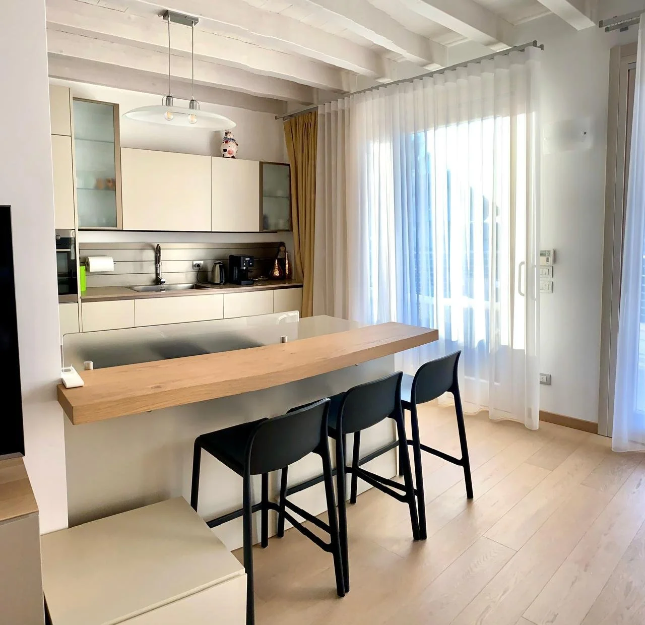 Modern kitchen with white cabinets, a black coffee maker, and a small breakfast bar with three black chairs. Large windows with sheer white curtains and yellow drapes provide natural light.