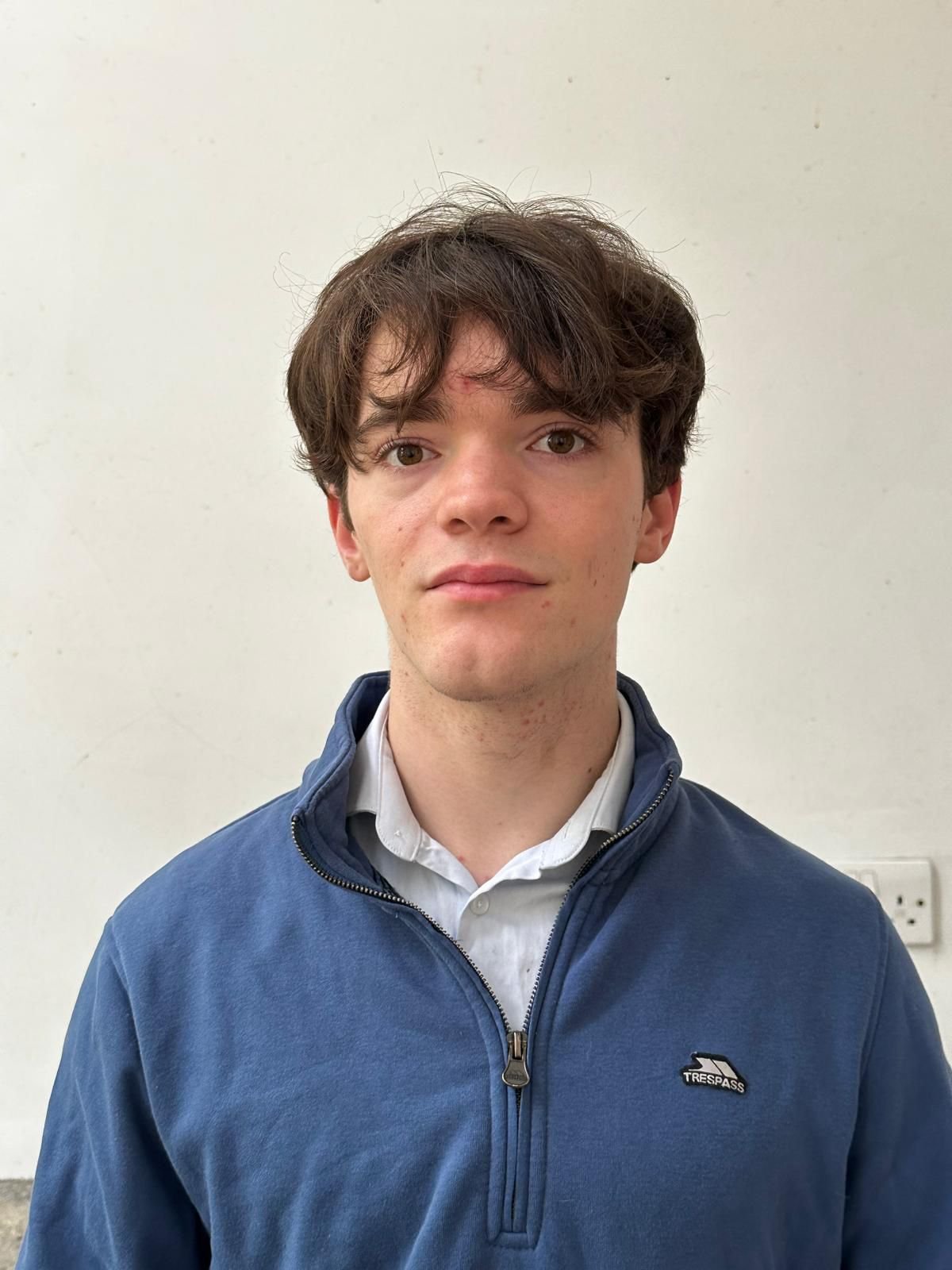 Portrait of a young man with brown hair and hazel eyes, wearing a white collared shirt and a blue zip-up jacket with a Trespass logo, standing against a plain white wall.