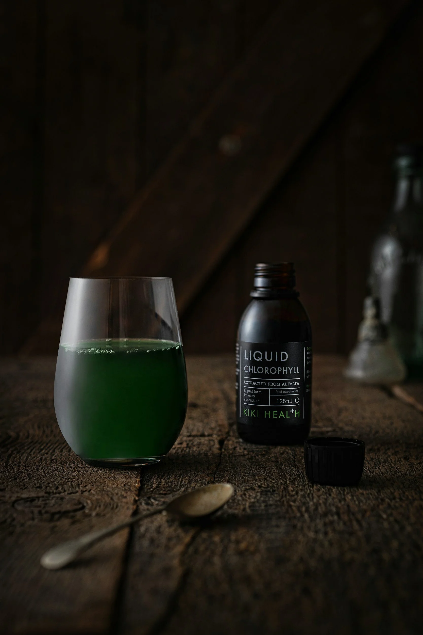 A glass of green liquid, a bottle labeled 'Liquid Chlorophyll,' and a spoon are on a wooden surface, with a dark background.