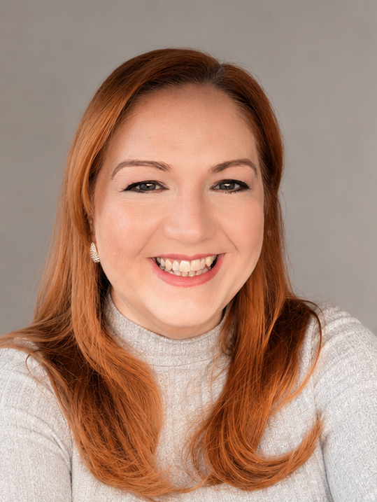 Close-up portrait of a smiling woman with red hair, wearing a light gray turtleneck sweater and pearl earrings, against a neutral background.