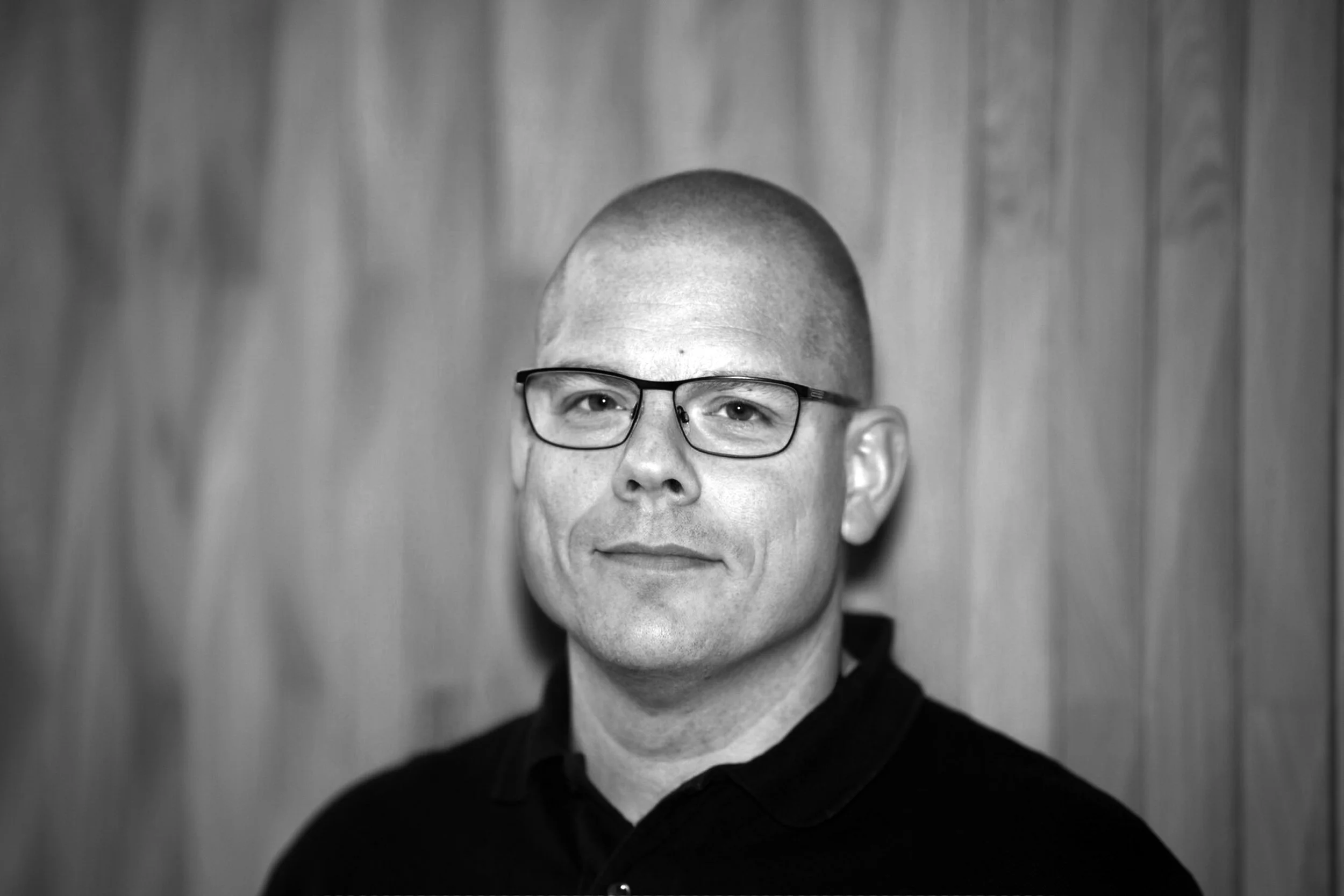 Schwarz-weiß Porträt eines Mannes mit Brille vor einer Holzpaneelwand