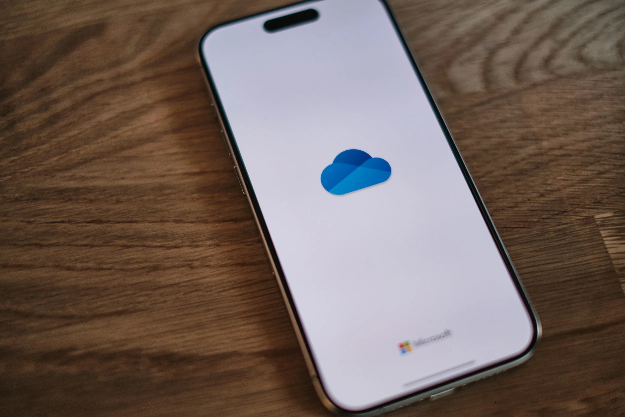 An iPhone lying on a wooden surface displaying the OneDrive app logo on its screen. Microsoft 365 cloud and Google Workspace conficuration and help.
