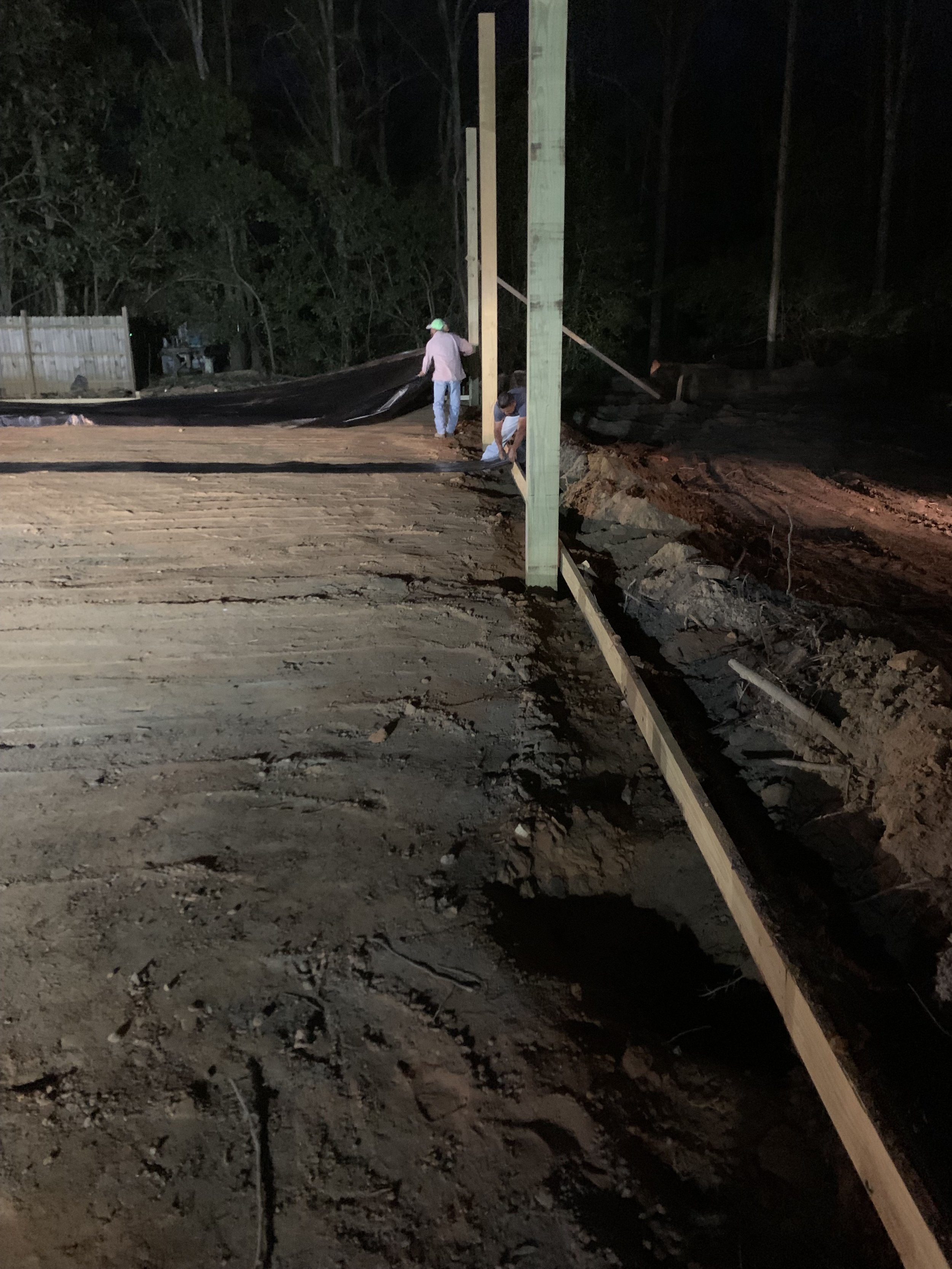 People working on a construction site at night, installing or repairing a wooden utility pole with a dirt ground and trees in the background.