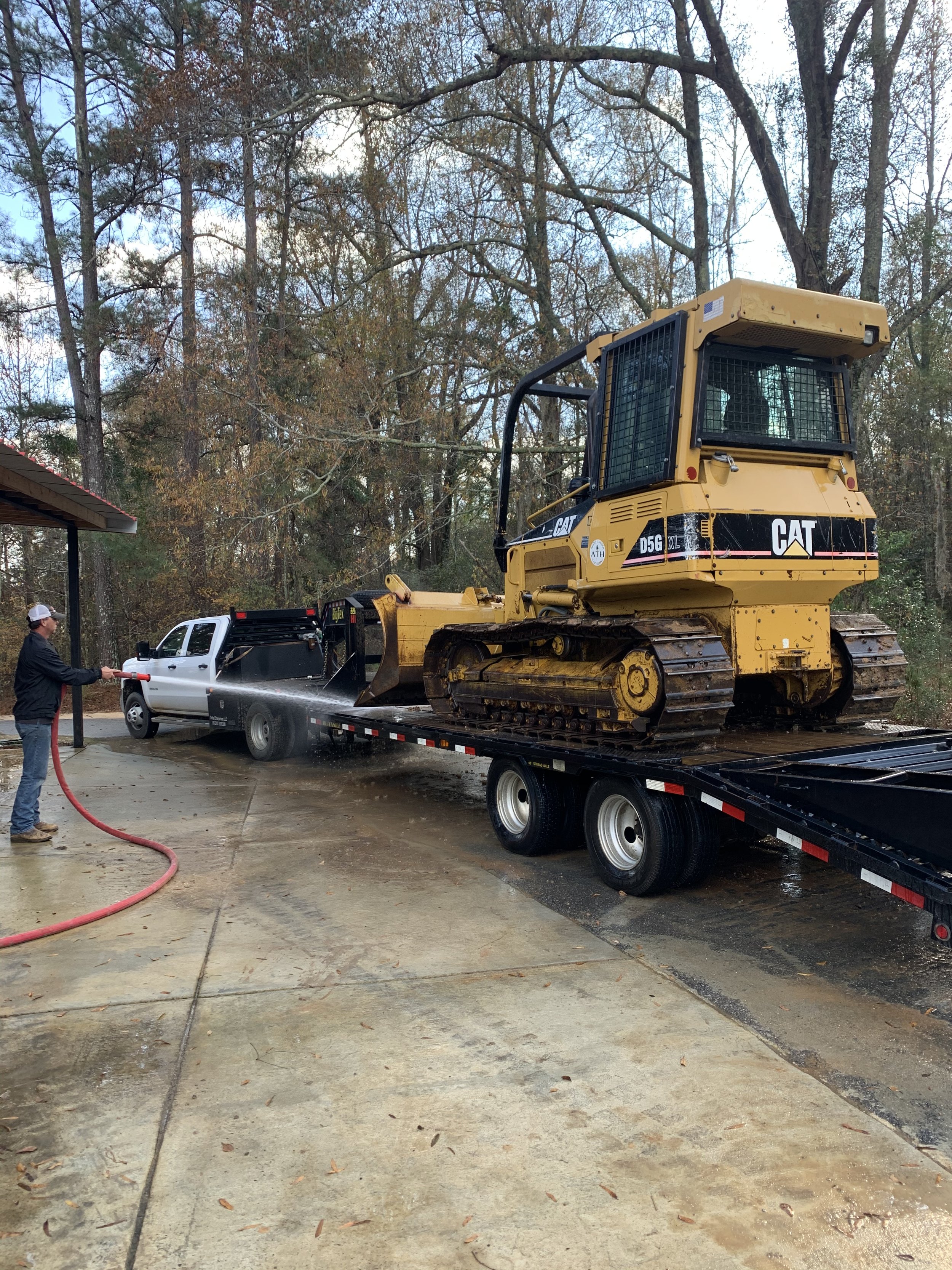 Person washing mud off a flatbed trailer carrying a yellow CAT bulldozer with a gray pickup truck in the background, trees, and a cloudy sky.