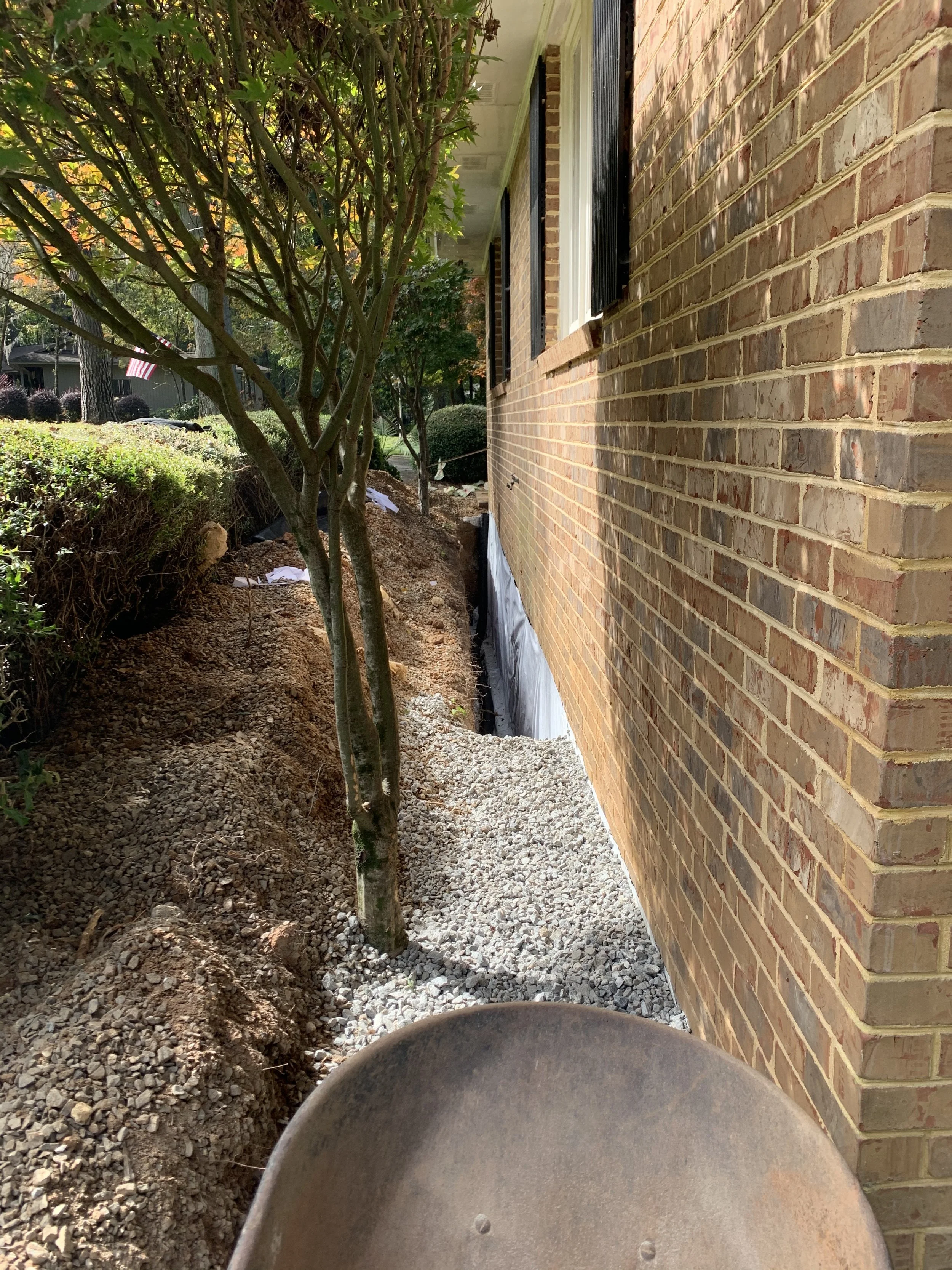 Side of a house with a brick wall, a window with black shutters, a tree with green leaves, and landscaping work in progress with soil and gravel.