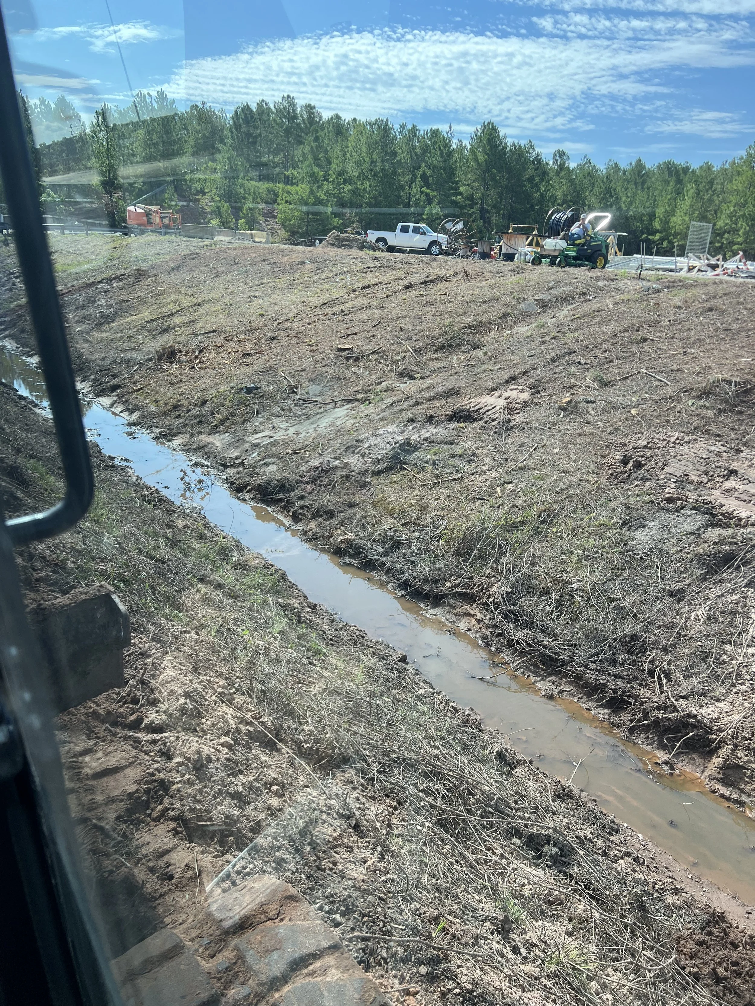 A small trench or creek running through a dirt area at a construction site, with a line of trees and construction vehicles in the background under a cloudy sky.