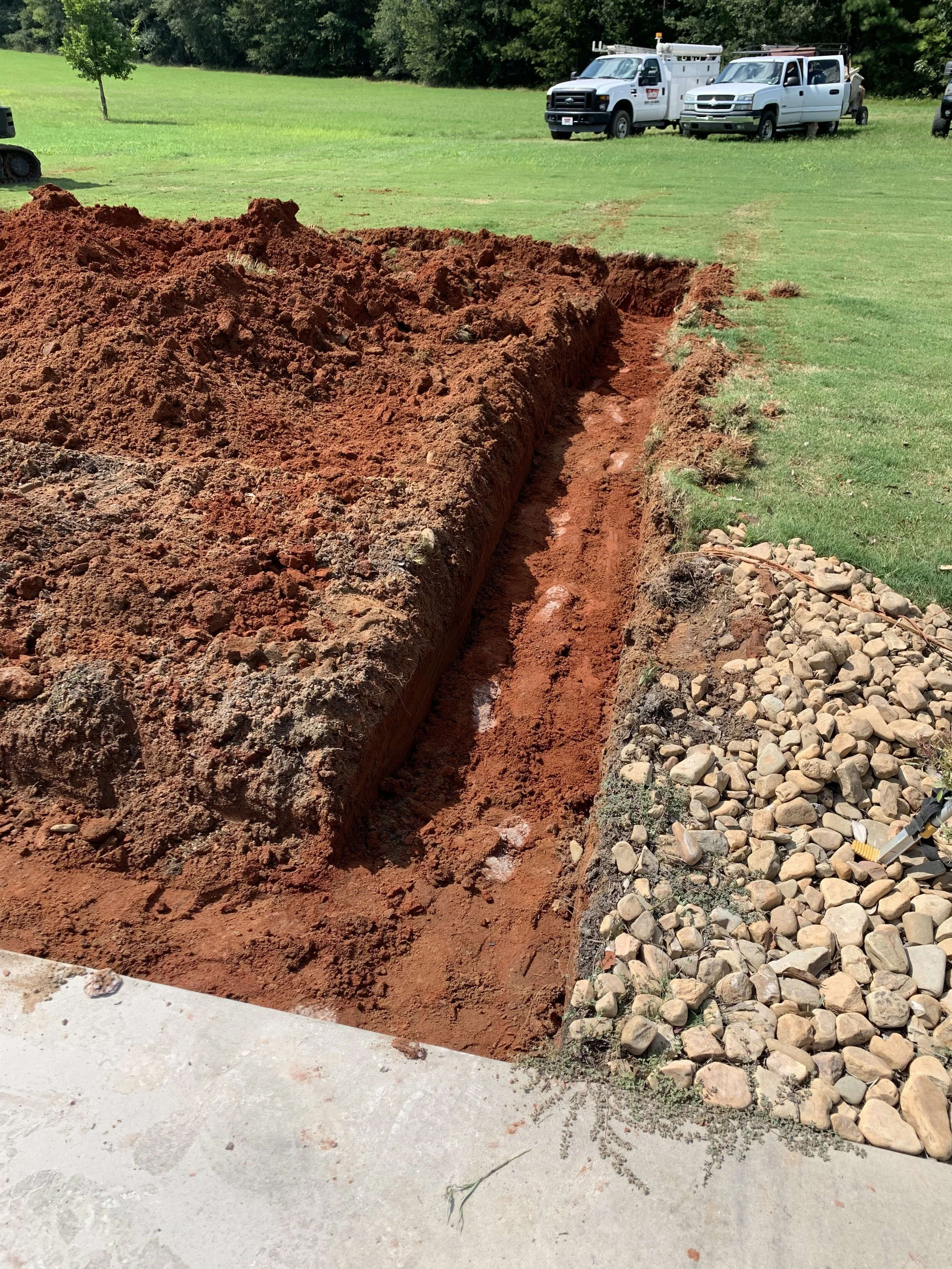 A trench is being dug in a grassy yard with piles of red dirt on one side and a strip of decorative rocks on the other. In the background, there are several utility trucks and a lawn mower.
