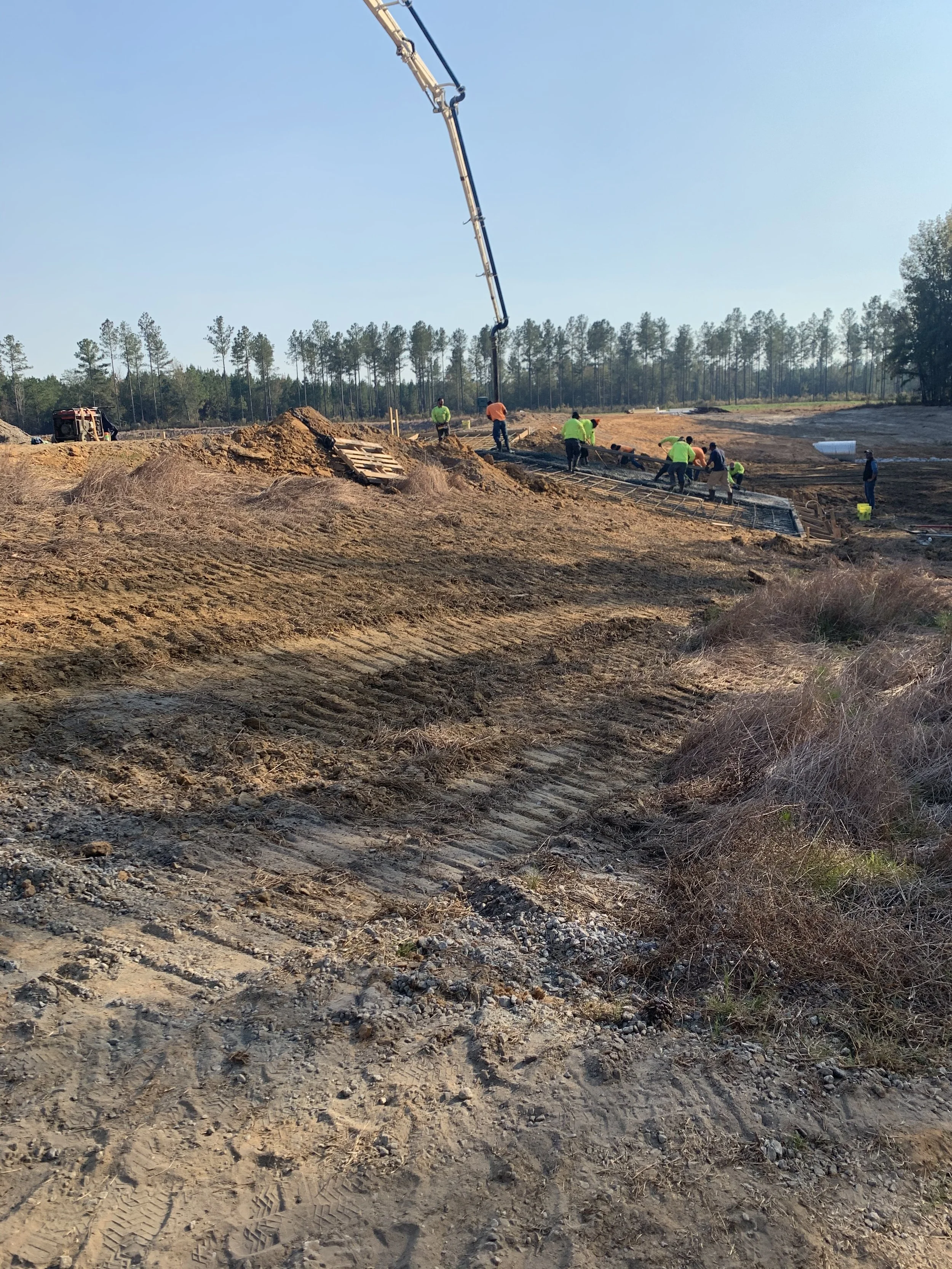 Construction workers pouring and leveling concrete on a construction site with a concrete pump truck and machinery in the background, surrounded by dirt and sparse vegetation.