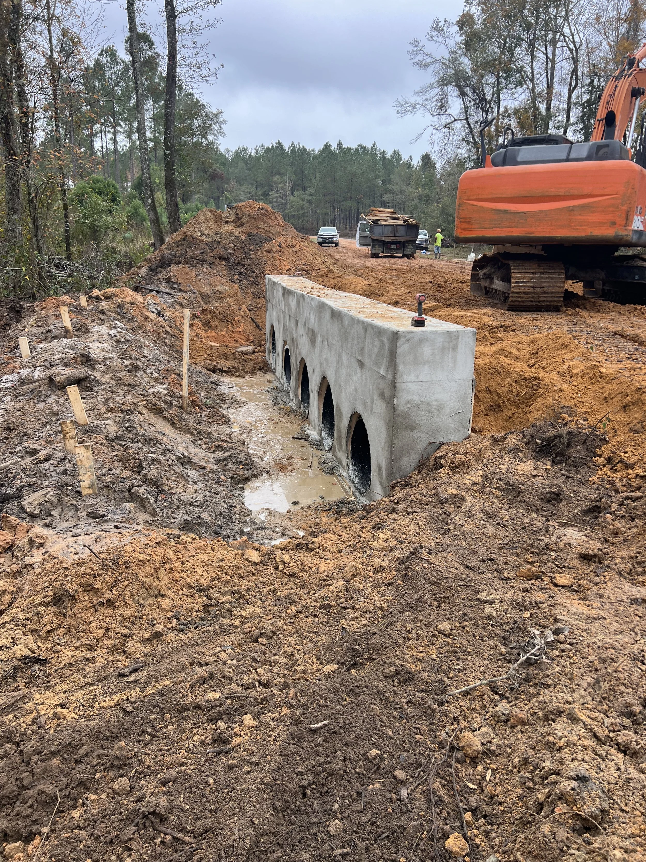 Construction site with a partially built concrete culvert with five arches, an orange excavator, dirt piles, and construction vehicles, with workers and trees in the background.