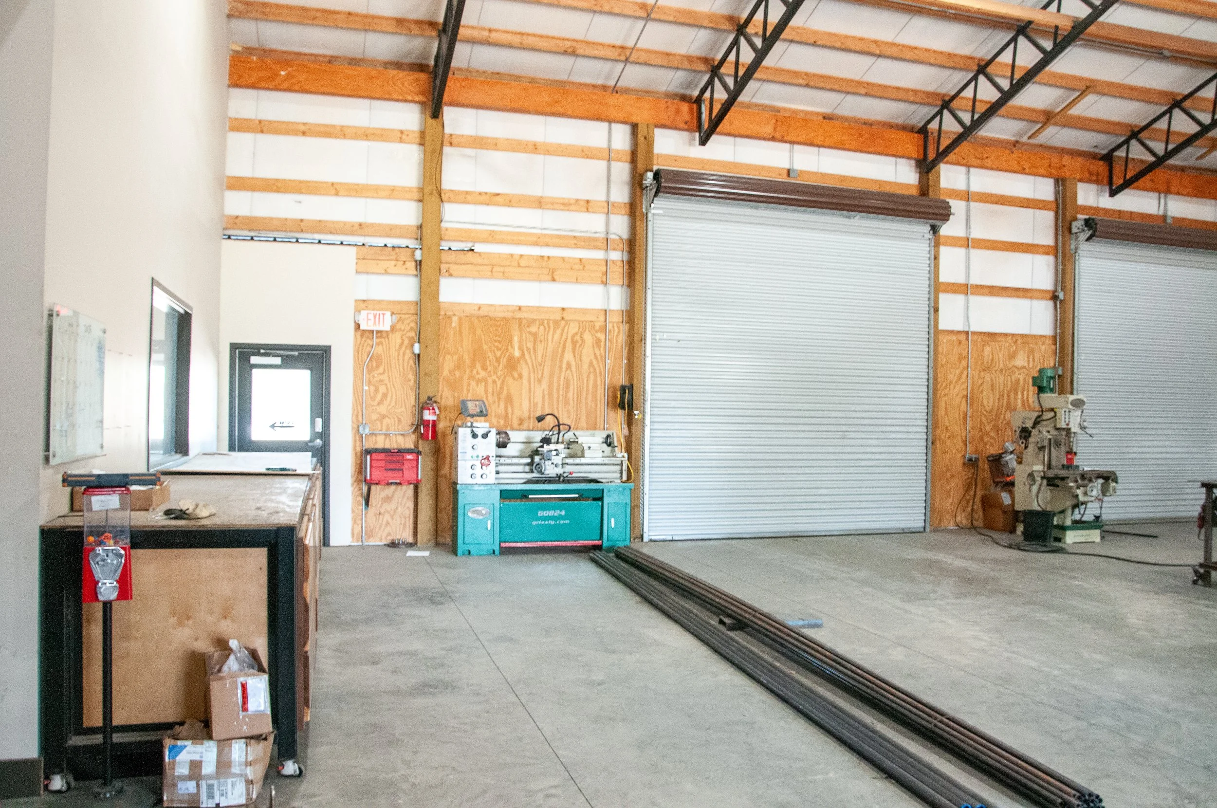 Interior view of an industrial workshop with a concrete floor, wooden wall panels, and metal roll-up doors. Equipment includes a blue machine, a vertical milling machine, a work table, and storage boxes.
