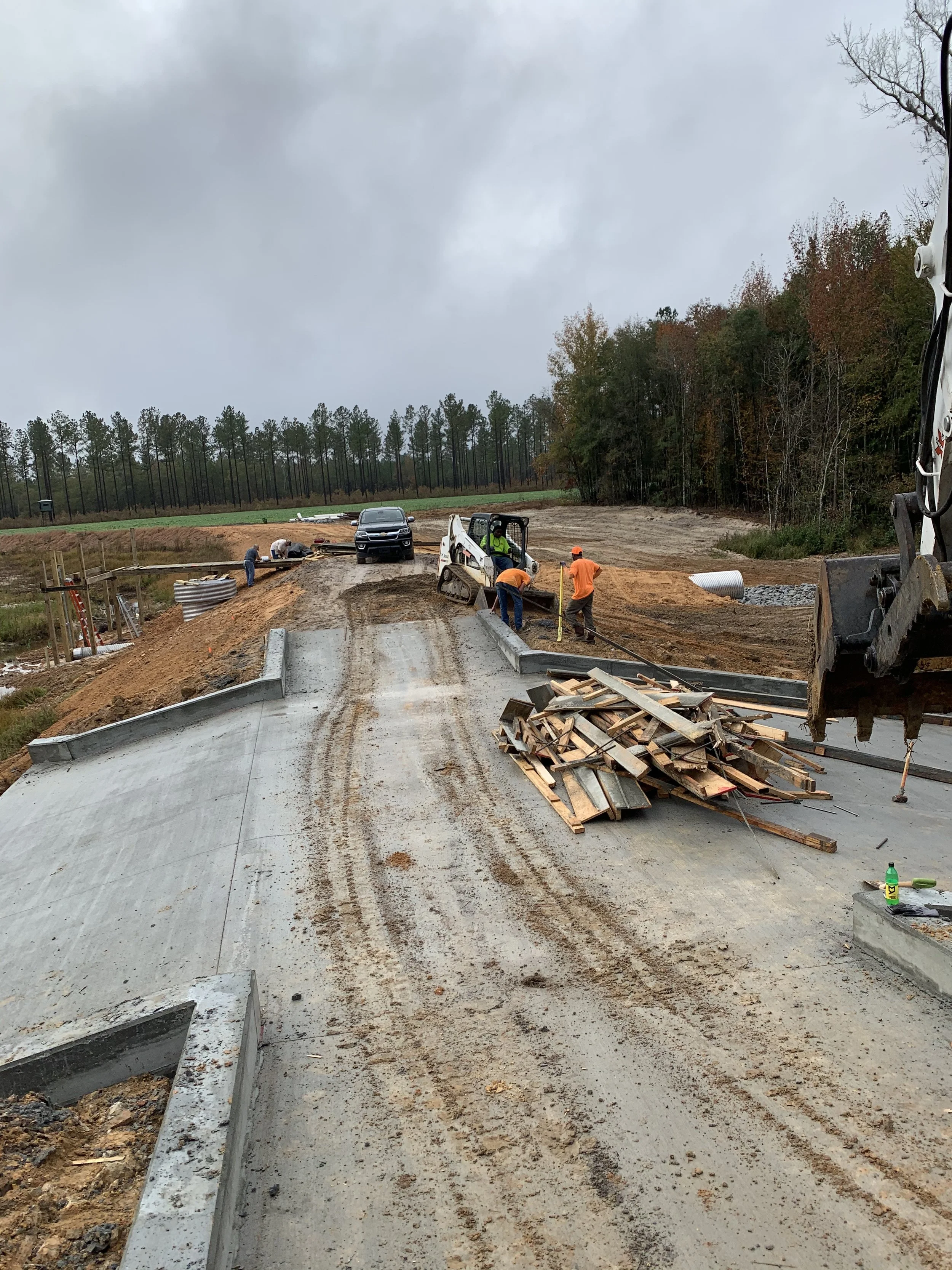 Construction workers building a road with concrete pavement, surrounded by construction materials and machinery, with a wooded area and cloudy sky in the background.