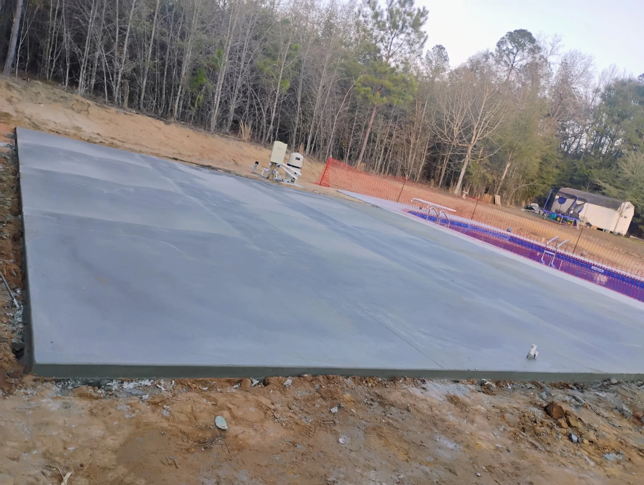Concrete pool deck foundation with underground plumbing in backyard, with trees and a shed in the background.