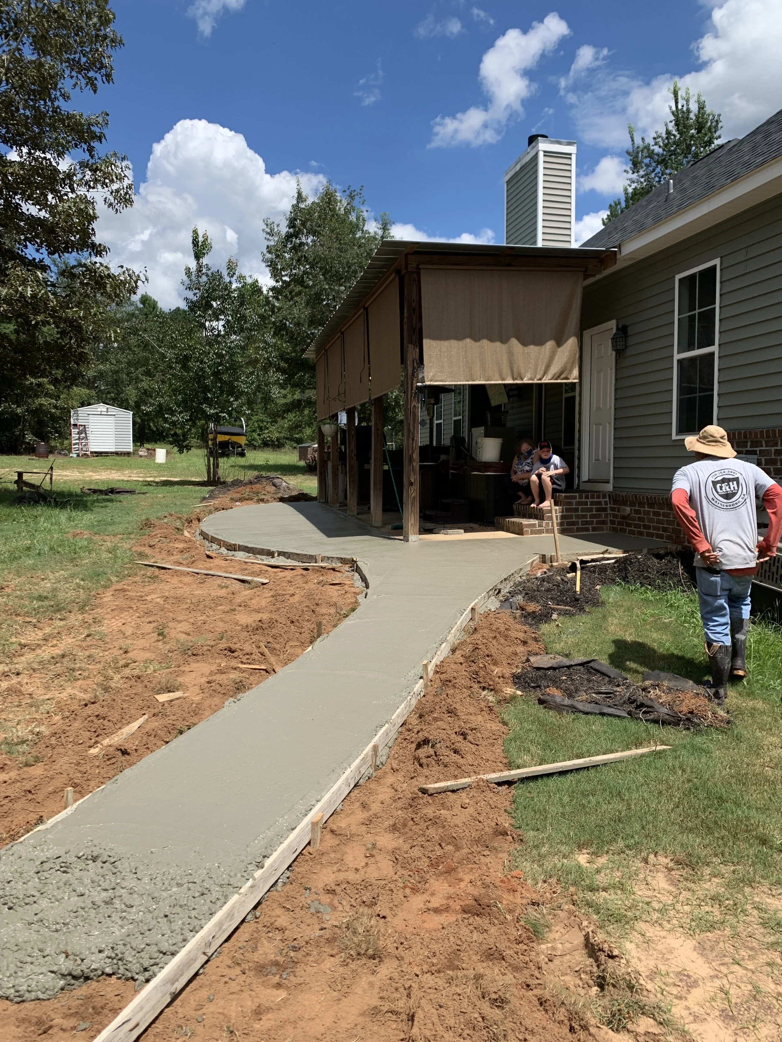 People working on the construction of a concrete walkway leading to a house porch under a partly cloudy sky.