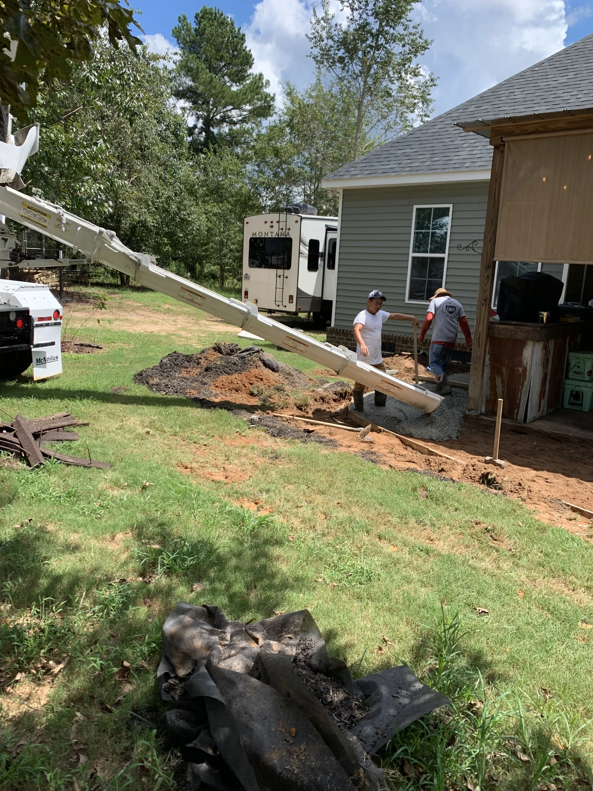 Workers pouring concrete for a sidewalk or patio at a residential construction site, with a concrete pump truck and a house with gray siding and white trim in the background.