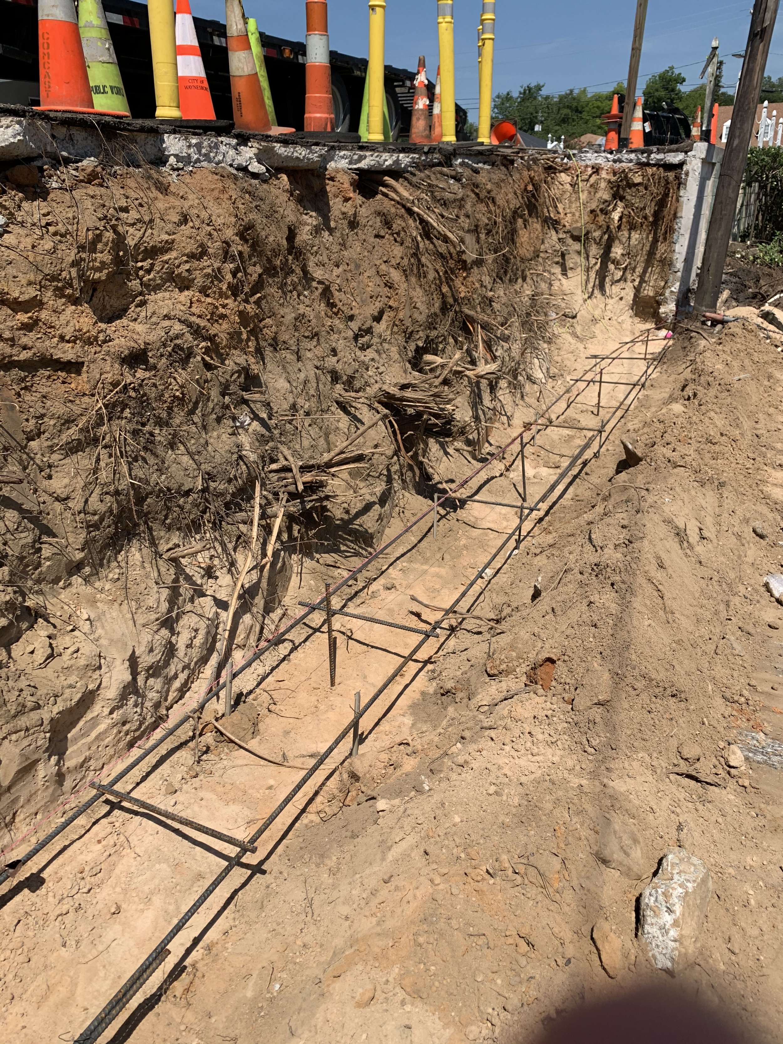 Construction site with exposed dirt and roots, with rebar set for foundation or retaining wall, and traffic cones along the edge.