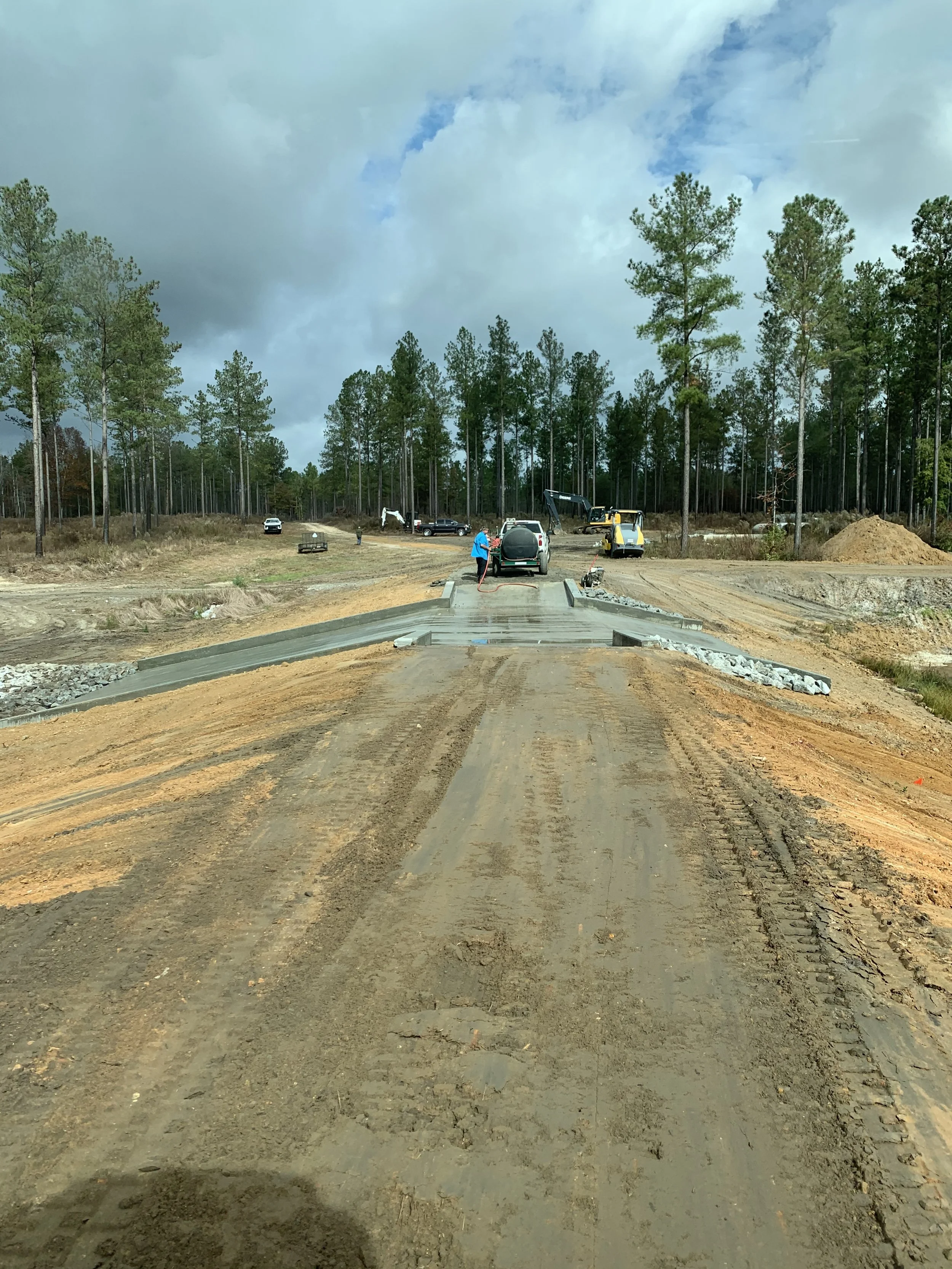 Construction workers building a concrete bridge on a dirt road in a forested area, with construction equipment and vehicles in the background.