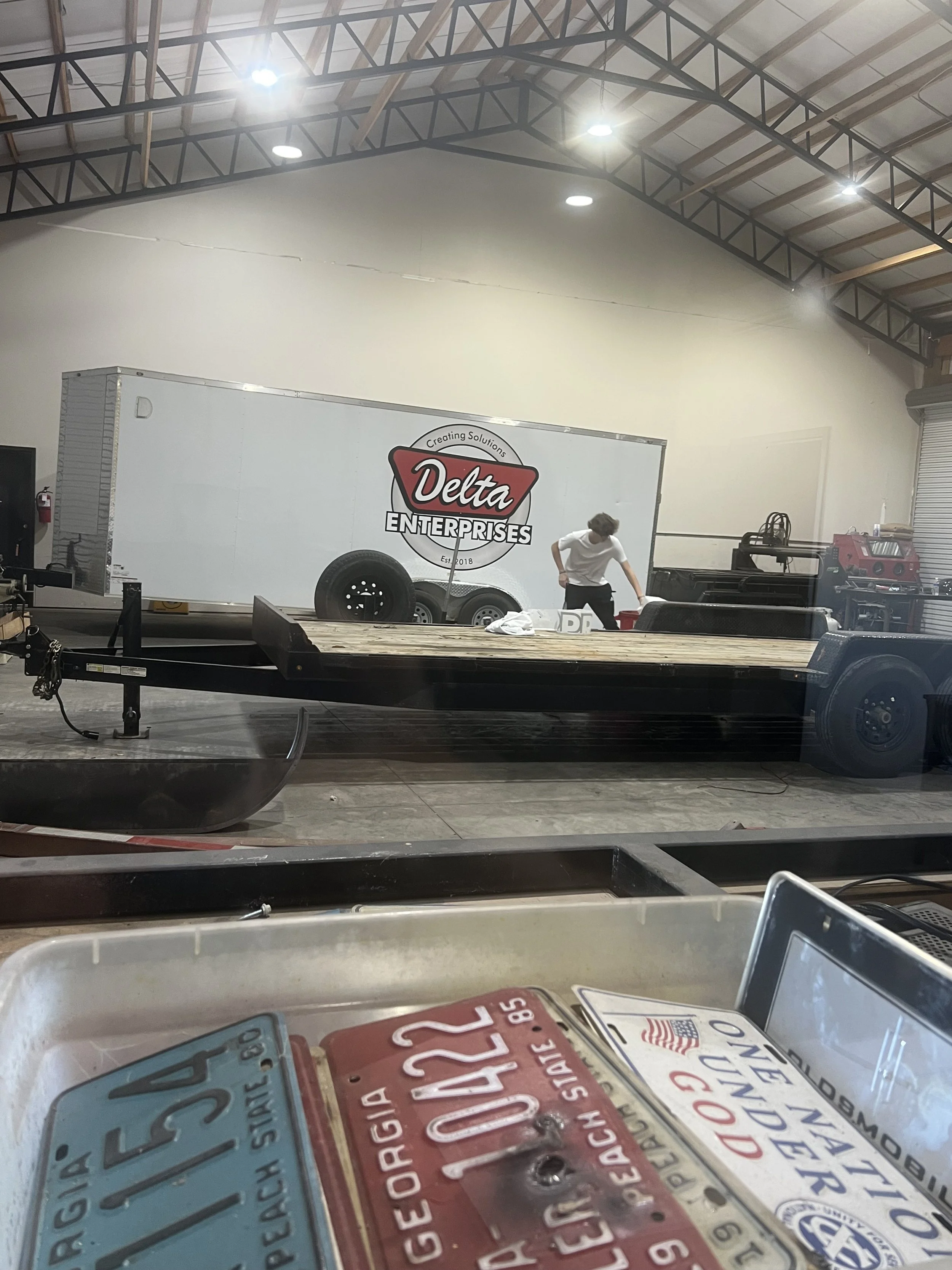 Inside a warehouse or workshop with a Delta Enterprises trailer, a person is working near various equipment, and there are some Georgia and Ohio license plates in the foreground.