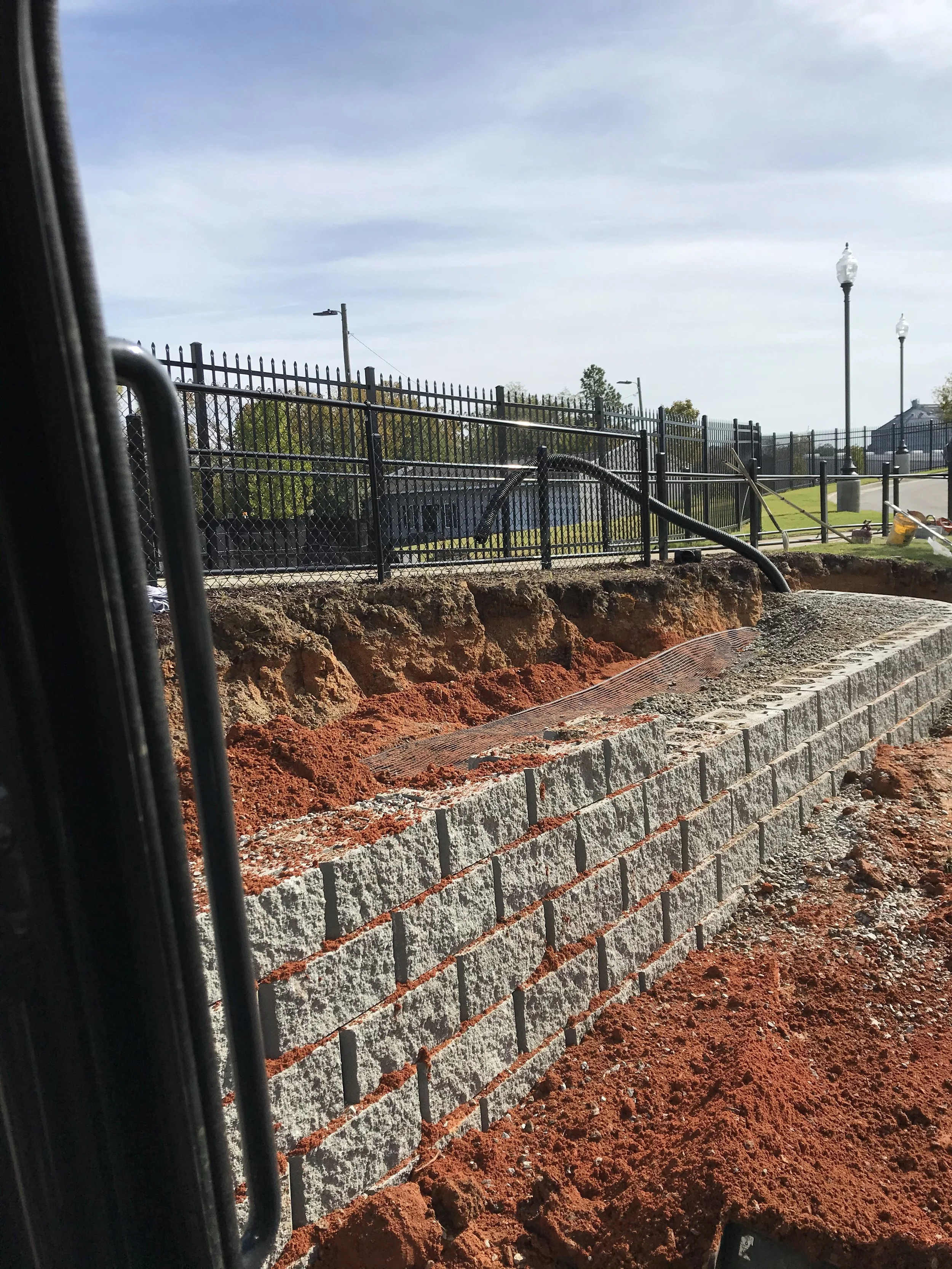 Construction site with a new brick wall being built, dirt and gravel in the foreground, black metal fence and street lamps in the background.