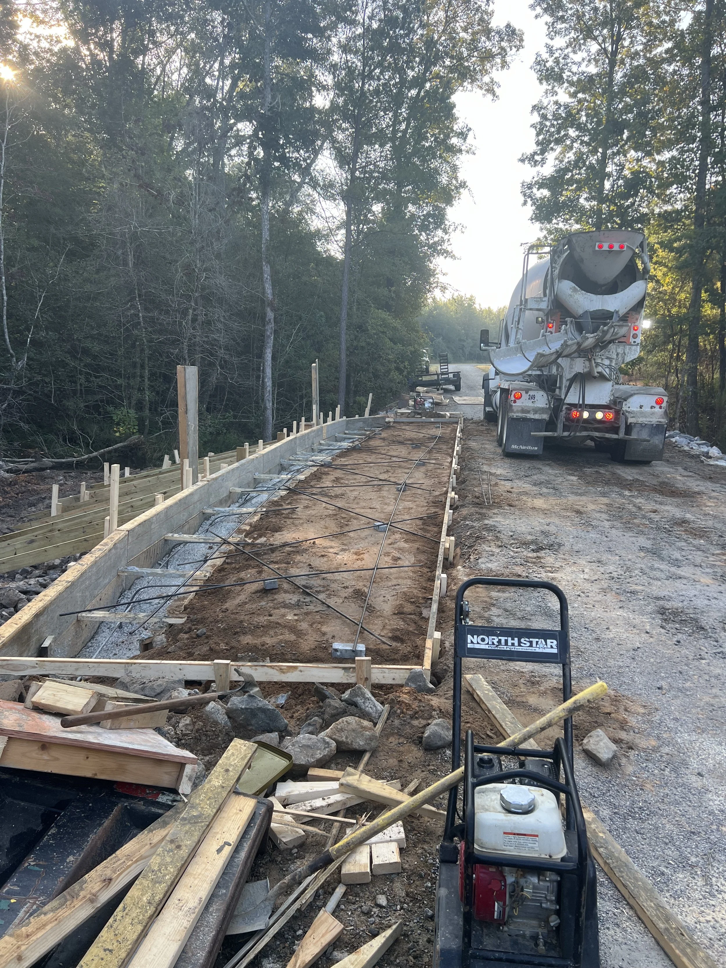 Construction site with a concrete mixer truck pouring concrete into a wooden frame for a pathway, surrounded by trees.