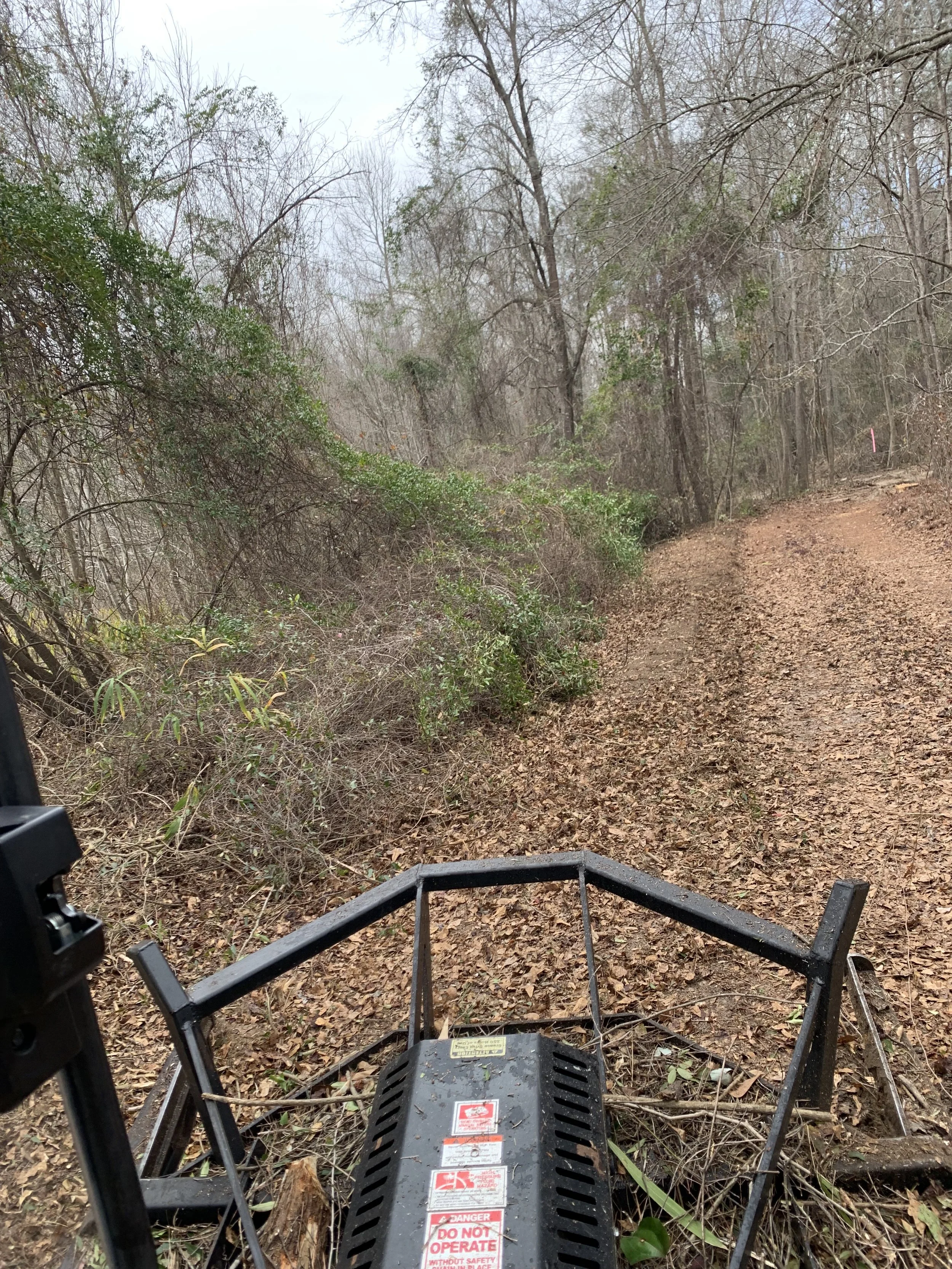 View of a dirt trail surrounded by trees and bushes, with an industrial machine at the bottom of the image.