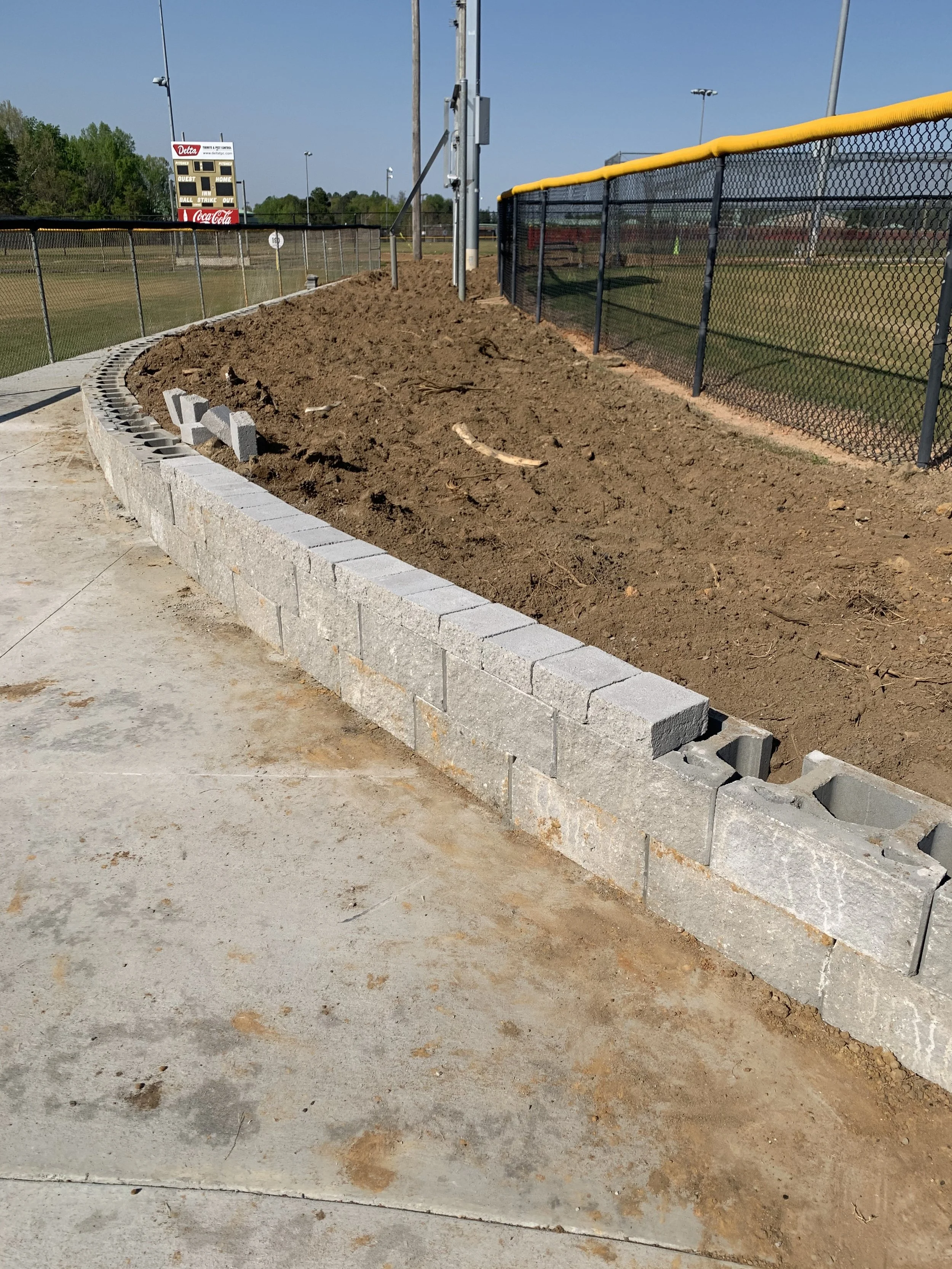 Partially built concrete block wall along a dirt raised bed at a sports field with a fence, scoreboard, and field lights in the background.