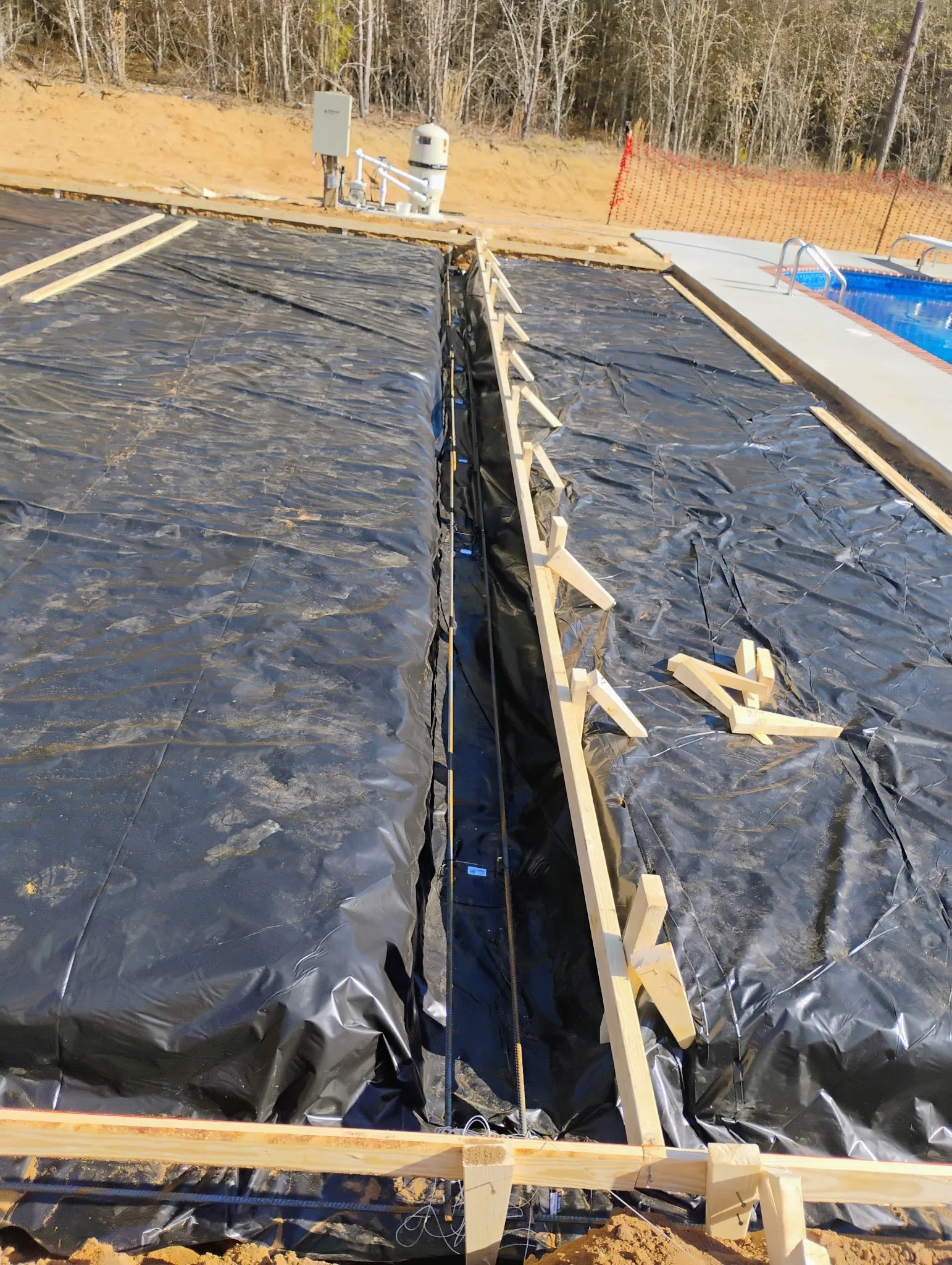 Construction site with black plastic sheeting, wooden stakes, and ropes, near a pool, with trees and orange safety fencing in the background.