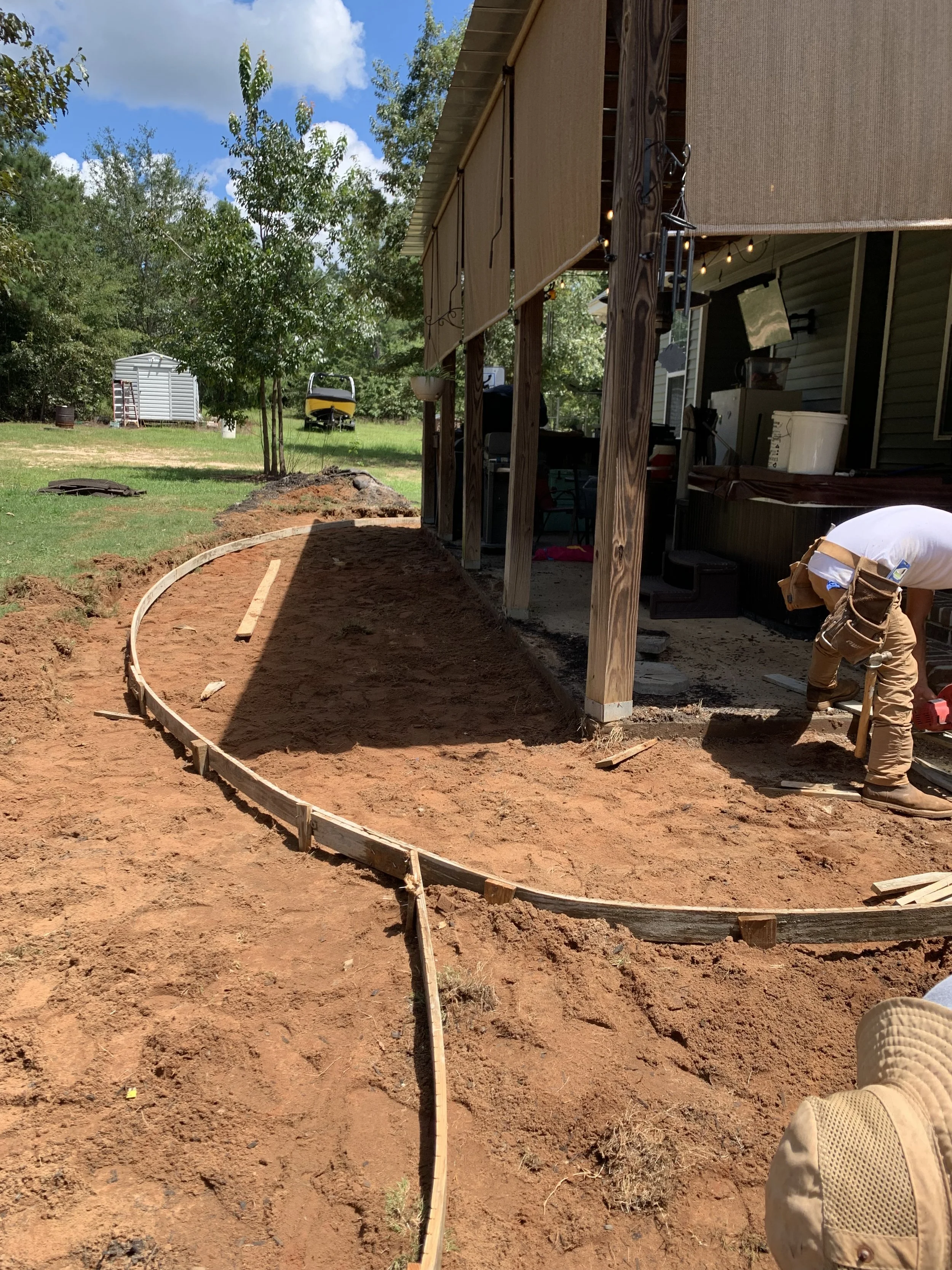 A construction worker is building a curved garden bed outline with wooden boards in the backyard of a house. The yard has trees, a small shed, and a boat in the background, with partly cloudy skies above.