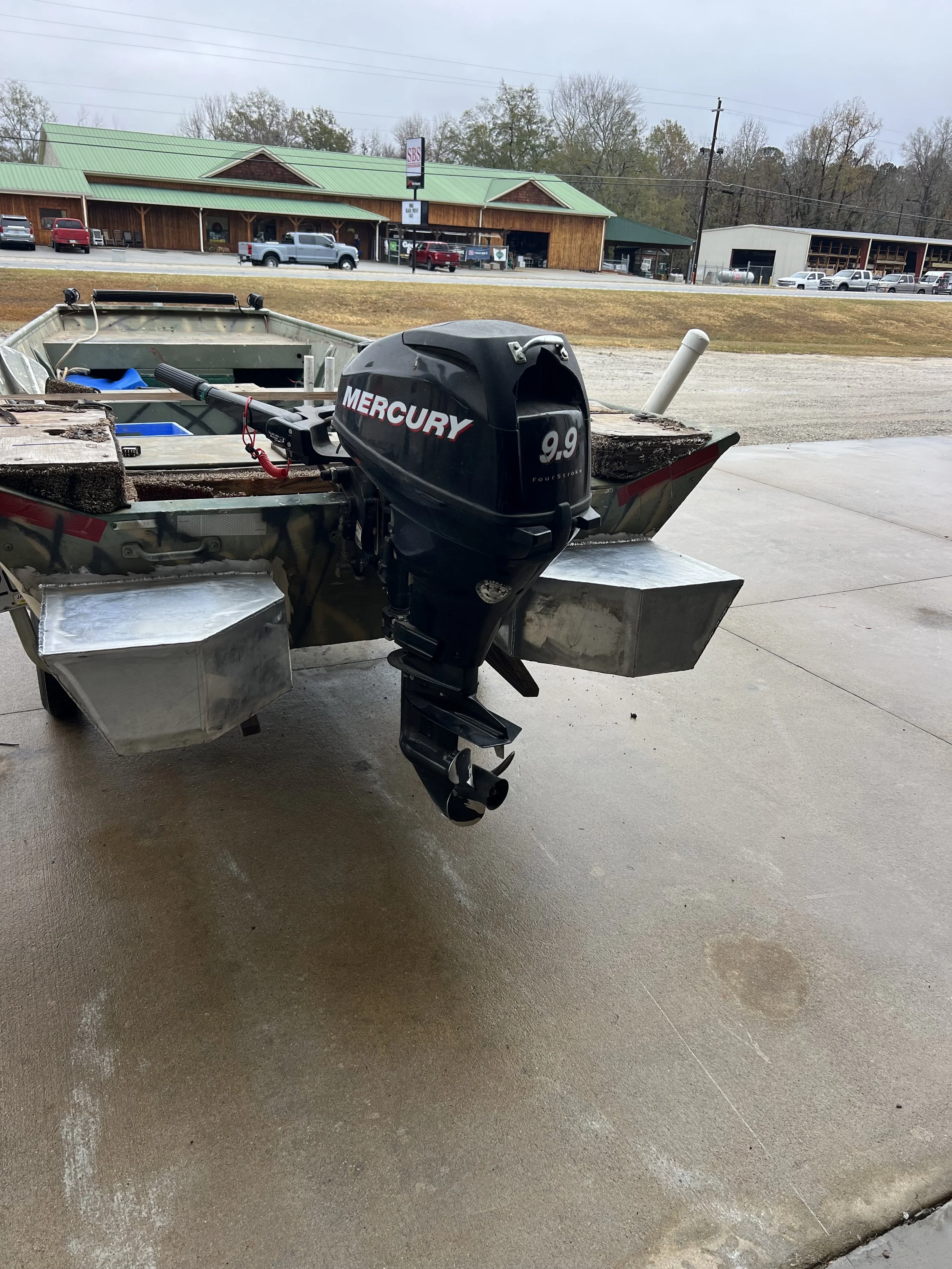 Small boat with a Mercury 9.9 horsepower outboard motor attached, parked on a concrete surface with a building and parked cars in the background.