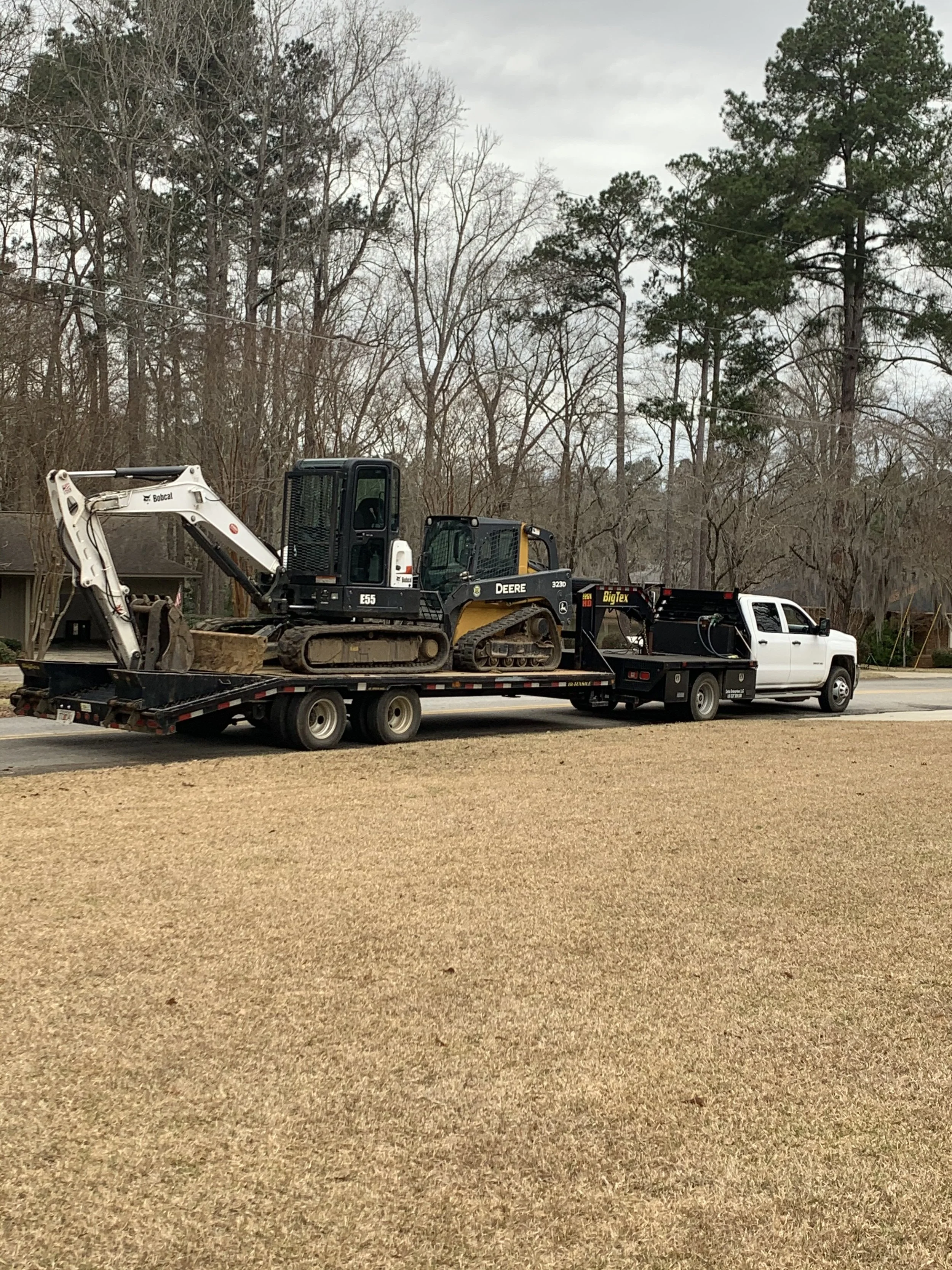 A flatbed towing truck carrying two construction vehicles, a small excavator and a bulldozer, on a residential street with leafless trees in the background.