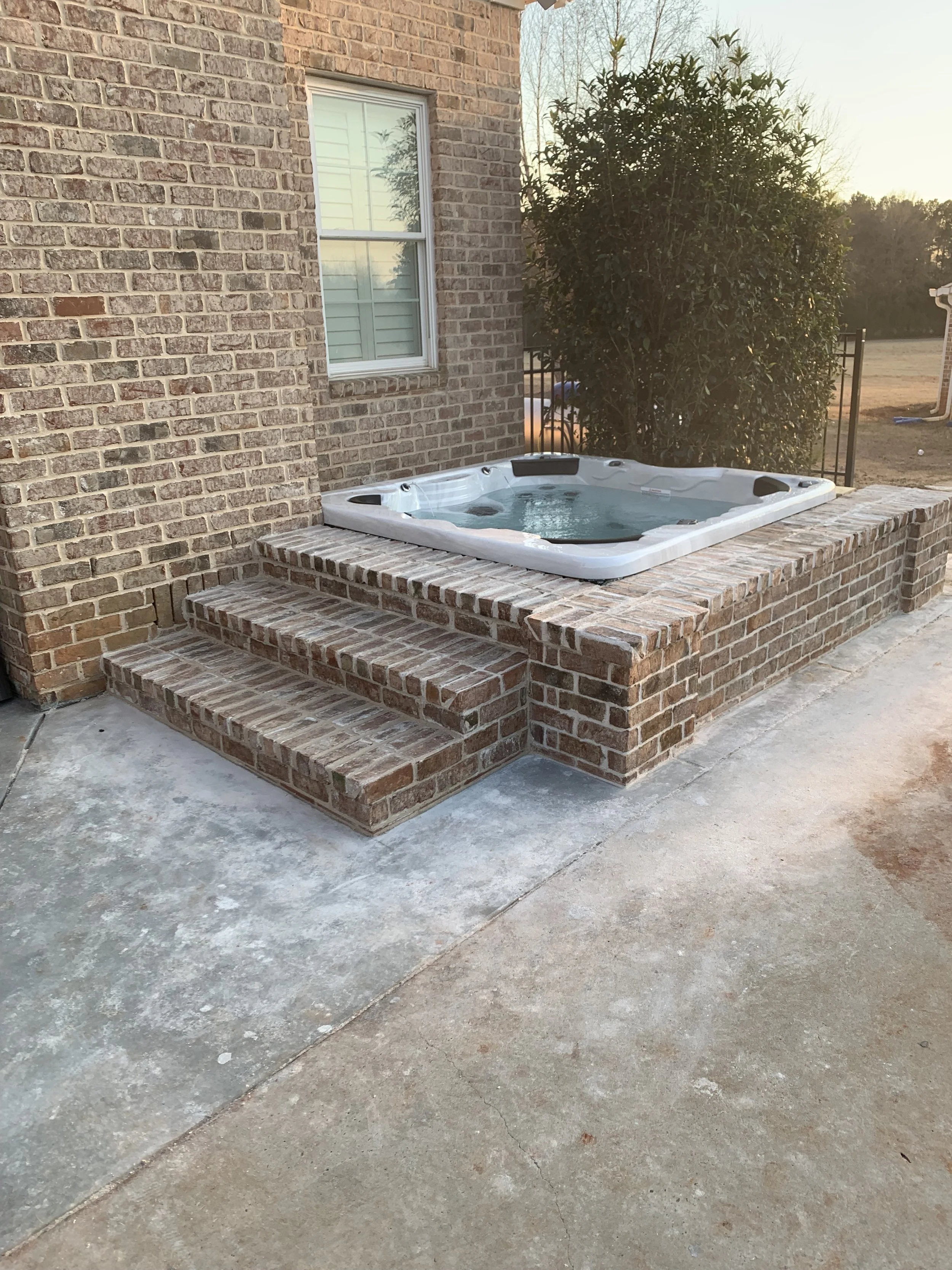A brick patio with a hot tub surrounded by a brick platform and brick steps, located next to a brick house with a window, during sunset.