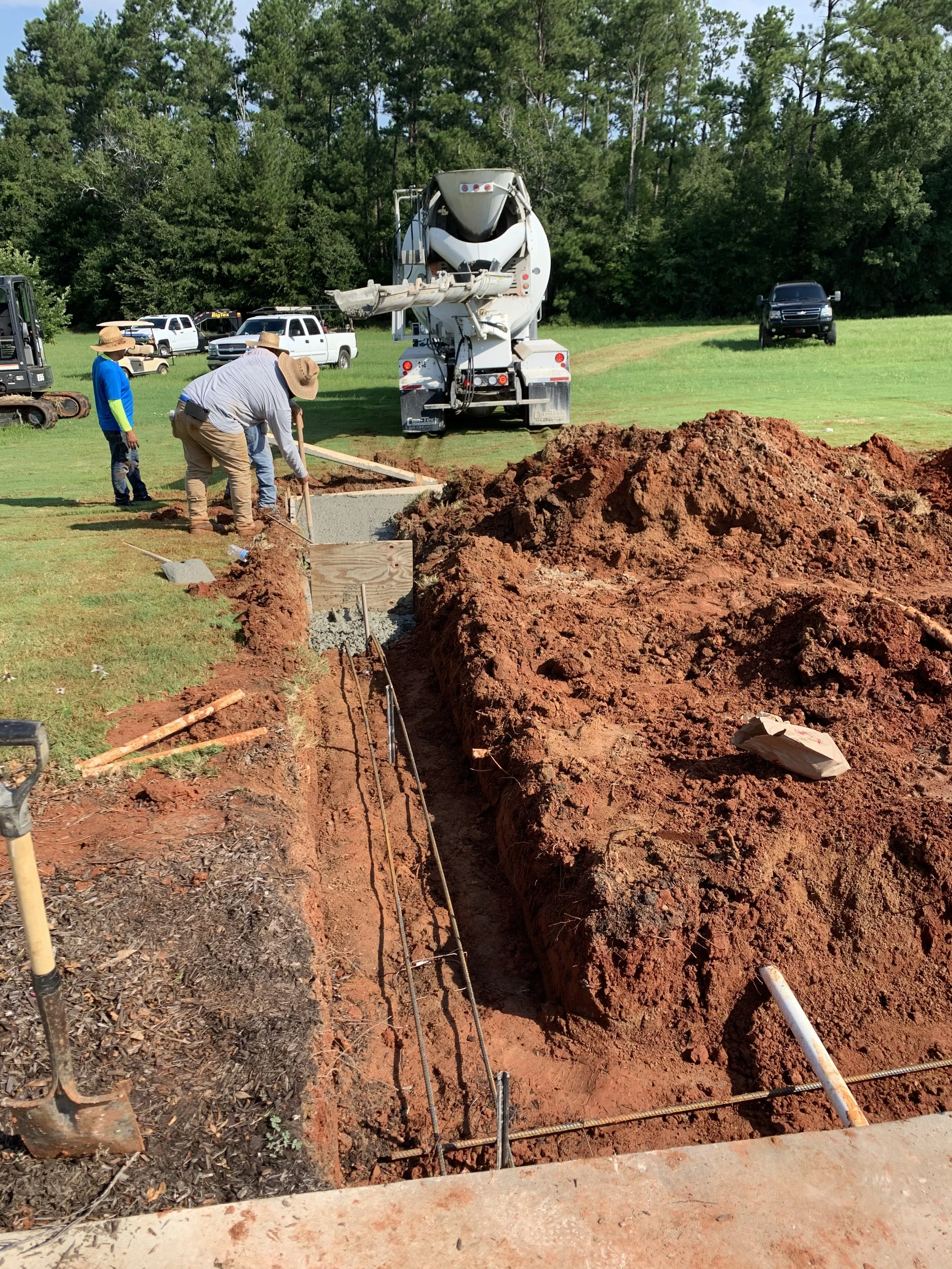 Construction workers building a foundation with a trench. A concrete mixer truck is present, with trucks and construction equipment in the background. The site is surrounded by trees and green grass.