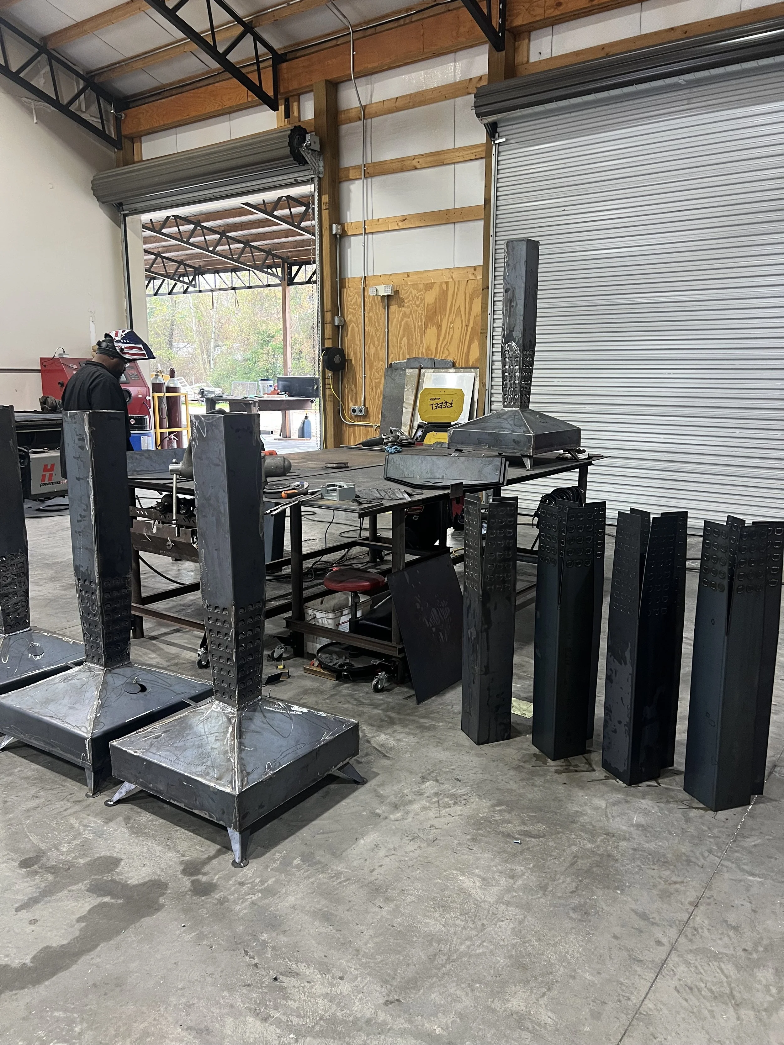 Industrial workshop with metal structures in progress, a person working, and a large open garage door showing outdoor trees and bright daylight.