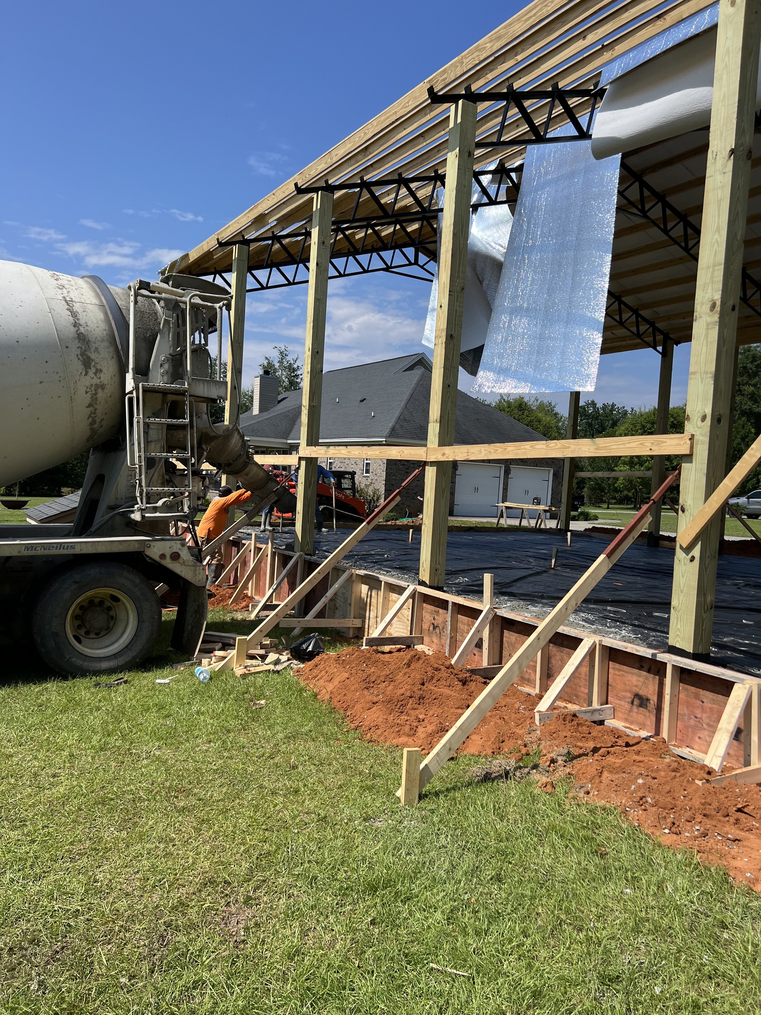 Construction site showing a foundation being prepared with a concrete mixer truck, wooden framework, and house in the background.